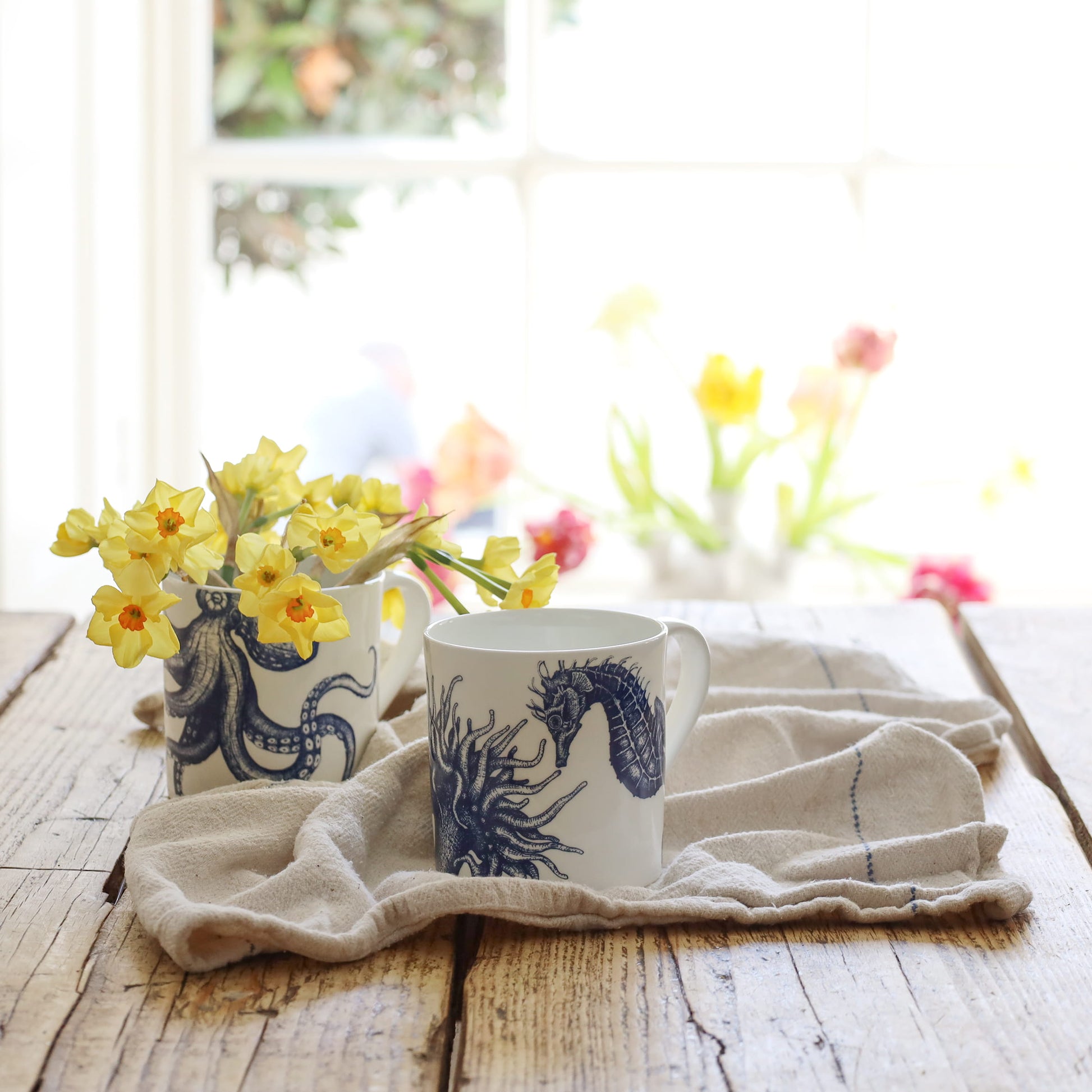 Two mugs with seahorse design on a wooden table with flowers in the background