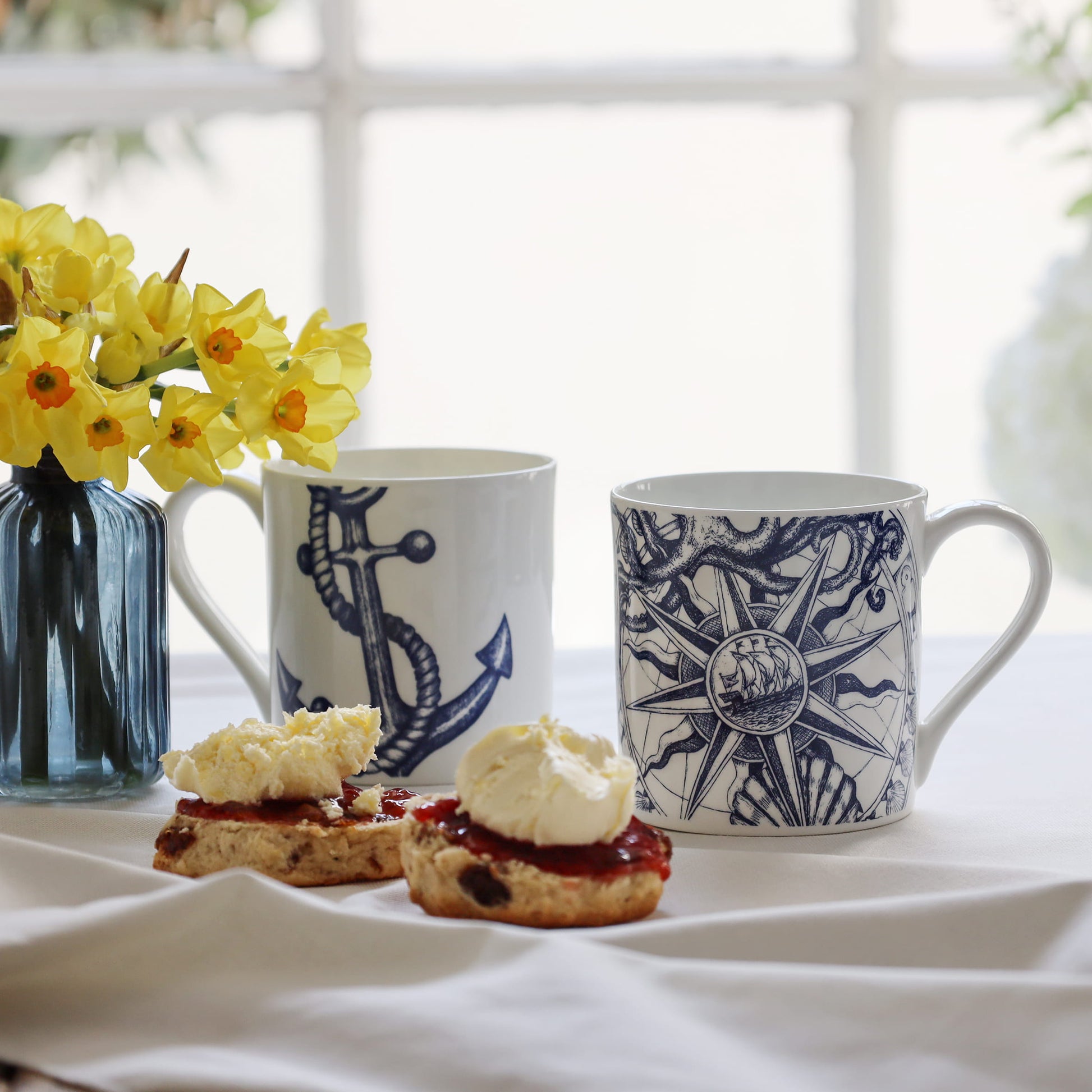 Two mugs with nautical designs, a vase of yellow flowers, and scones with cream and jam on a white surface.