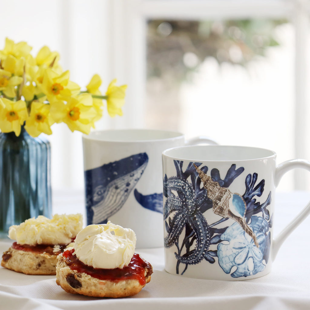 Two mugs with blue and white designs, accompanied by scones with cream and jam, on a light surface.