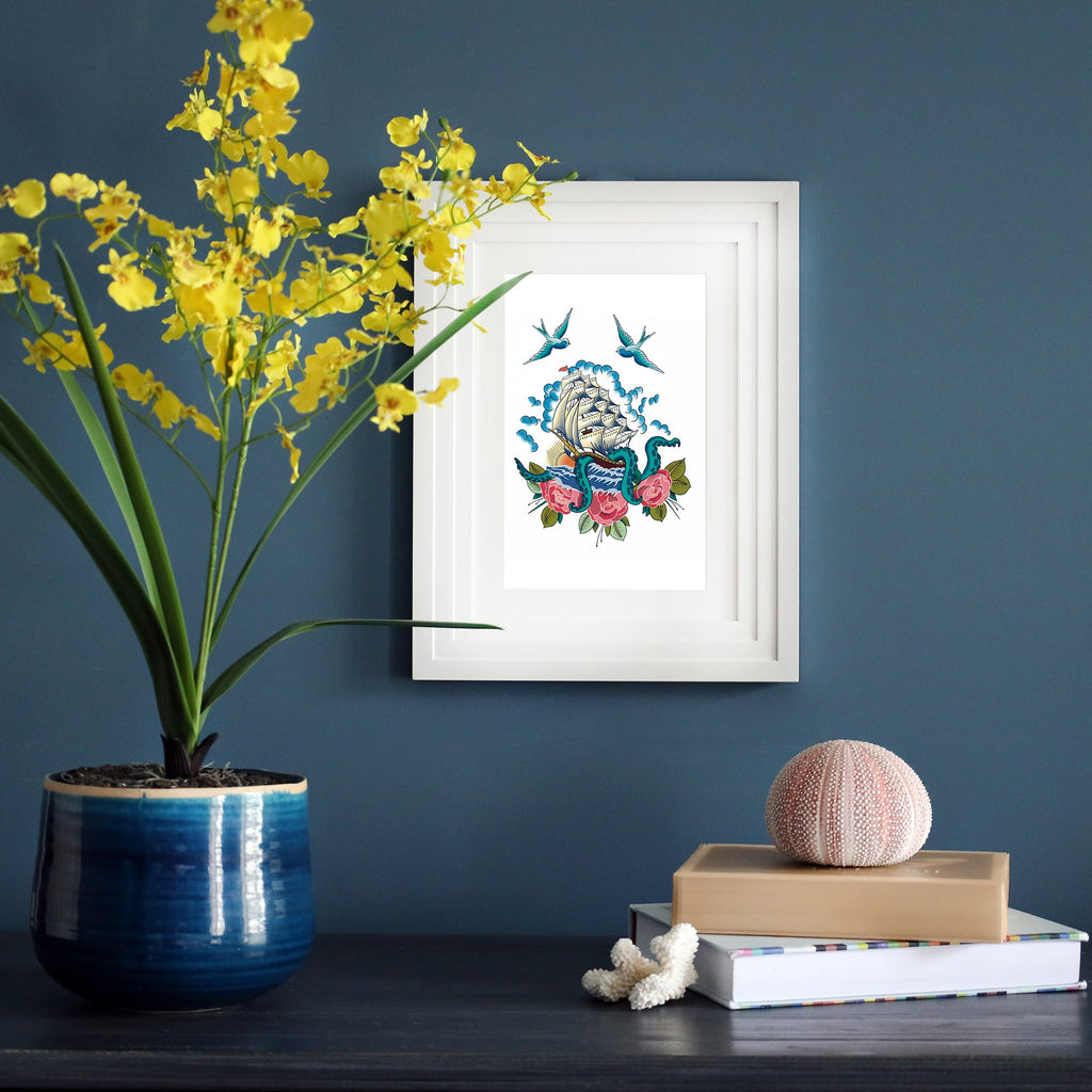 A framed nautical print on a dark blue wall, featuring a tattoo-style illustration of a sailing ship entangled with a kraken, surrounded by swallows and roses. The artwork is displayed on a stylish shelf with decorative elements, including a blue ceramic vase with yellow flowers, stacked books, and a sea urchin shell.