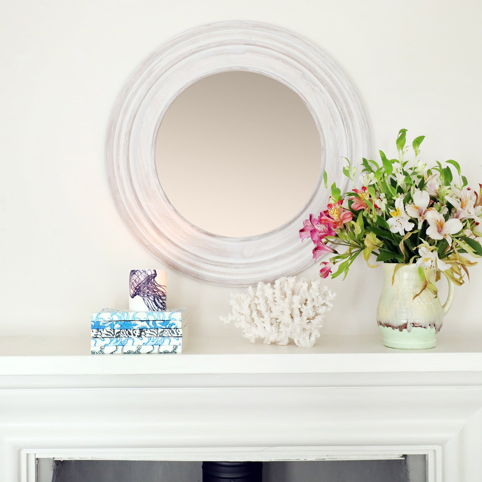 The image shows a white, circular mirror with a distressed frame, mounted above a white mantel. On the mantel, there is a green ceramic pitcher filled with a bouquet of fresh flowers in shades of pink, white, and green. To the left of the pitcher is a piece of white coral, and beside it, there are two small decorative items: a blue and white box and a small candleholder with a jellyfish design. The arrangement has a clean, coastal-inspired aesthetic, with a mix of natural elements and soft colors.