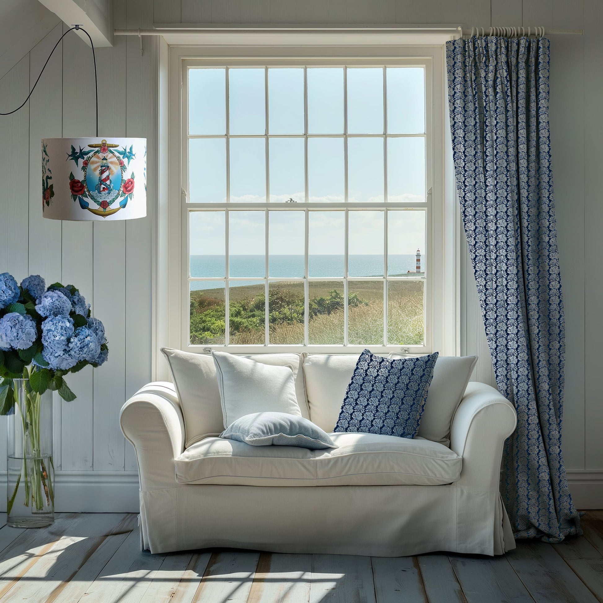 A serene coastal living room featuring a white sofa with soft blue and navy cushions, one of which has a detailed coral-inspired print. Matching navy curtains frame a large window that offers a stunning view of a lighthouse and the sea beyond. A bold, nautical-style lampshade and a vase of blue hydrangeas add to the seaside charm of the light-filled room.