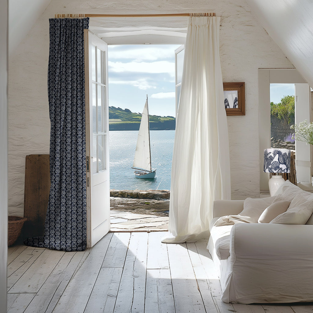 Coastal-inspired interior with an open French door revealing a sailboat on calm water. The room features white wooden floors, a cream sofa with neutral cushions, and curtains—one in sheer white and the other in a navy blue coral print. A nautical-themed lampshade and framed art complement the seaside aesthetic. Natural light fills the space, creating a serene and breezy atmosphere.