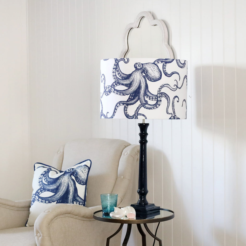 Navy Lacquer Godolphin lamp base with an octopus lampshade on a mirror table.On the table is a glass with some shells placed on it.Next to the table is a chair with an octopus cushion.Behind on the wall is a wave mirror.