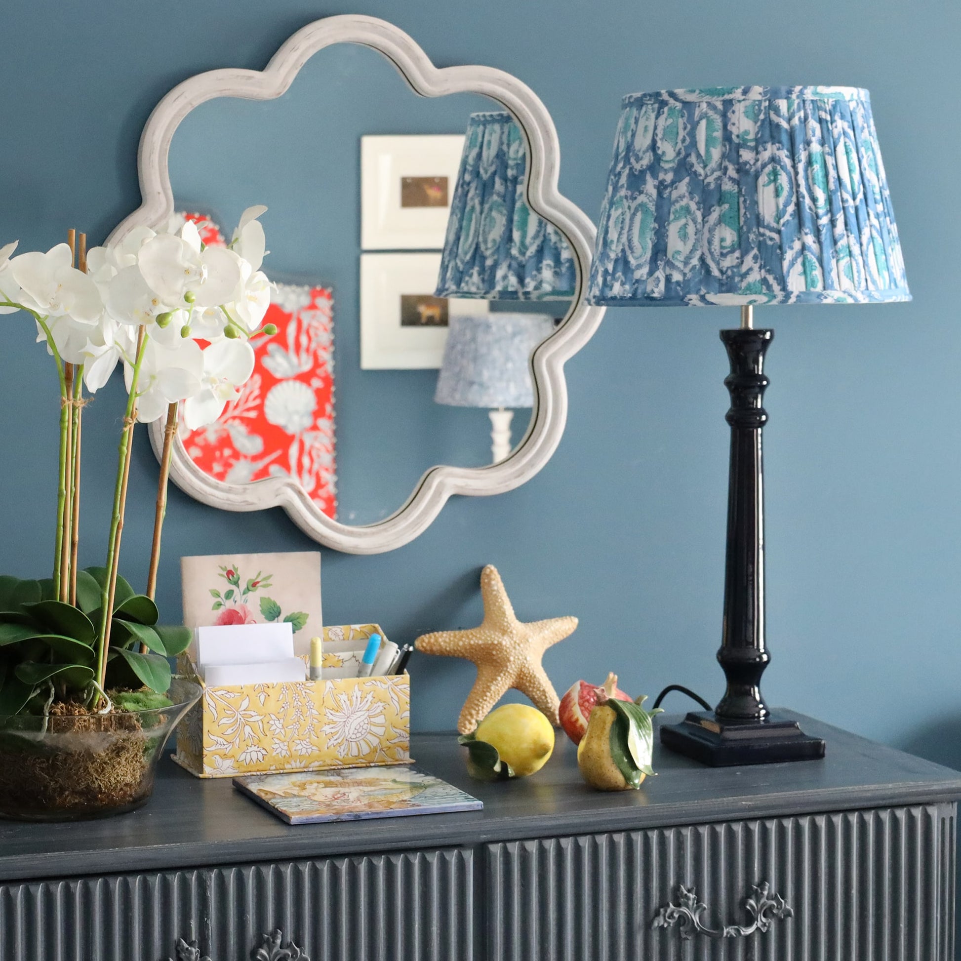 Whitewashed wooden wavey edged shaped mirror on the wall.In front is a sideboard with an orchid plant,some stationery, ceramic fruit and one of our navy lamp bases and a pleated lampshade