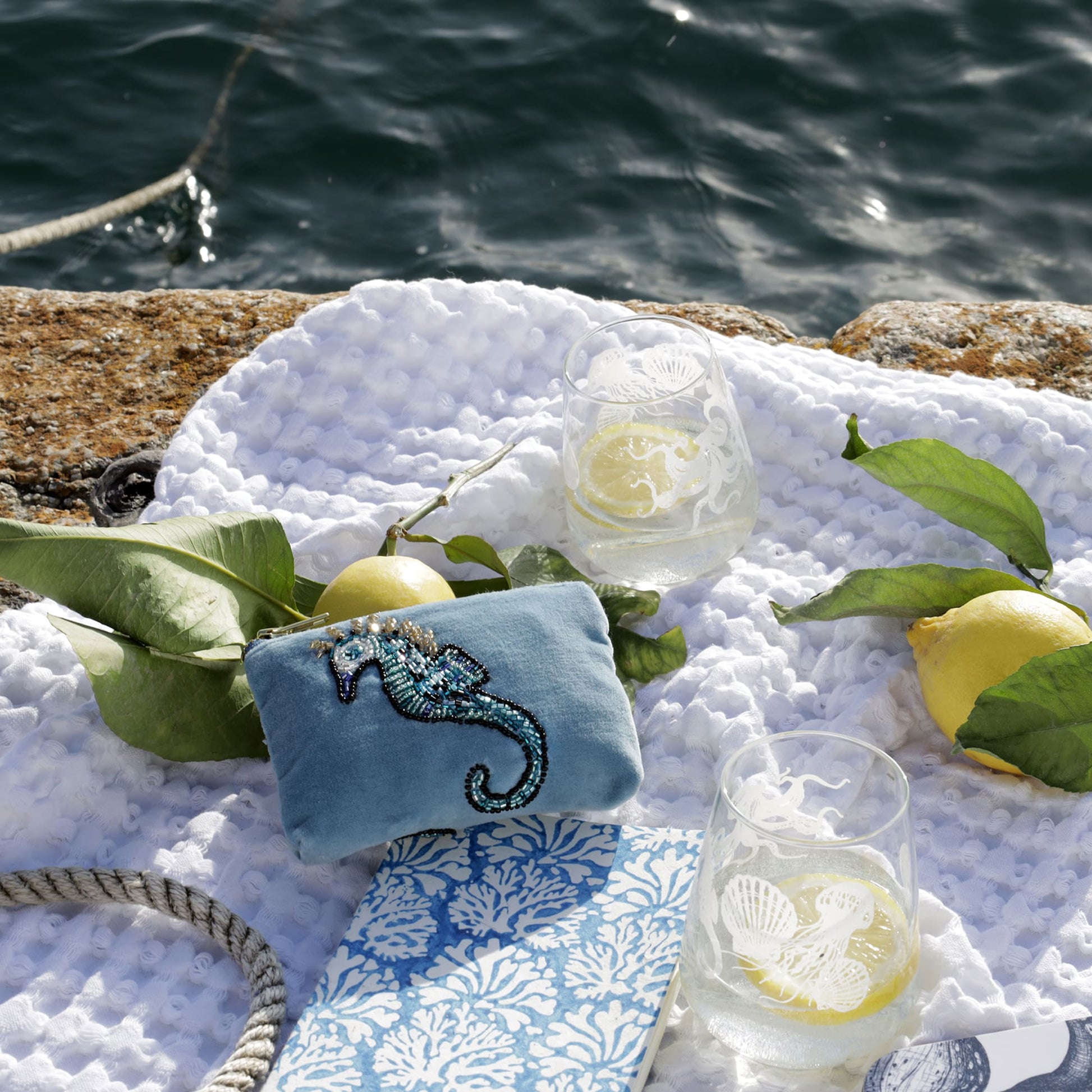 A small pale blue velvet purse with a beaded seahorse design is displayed on a textured white blanket by the seaside. Surrounding it are lemons with leaves, etched glasses filled with lemon water, a light blue coral-patterned notebook, and a section of nautical rope. The ocean glistens in the background, creating a breezy and refreshing coastal vibe.