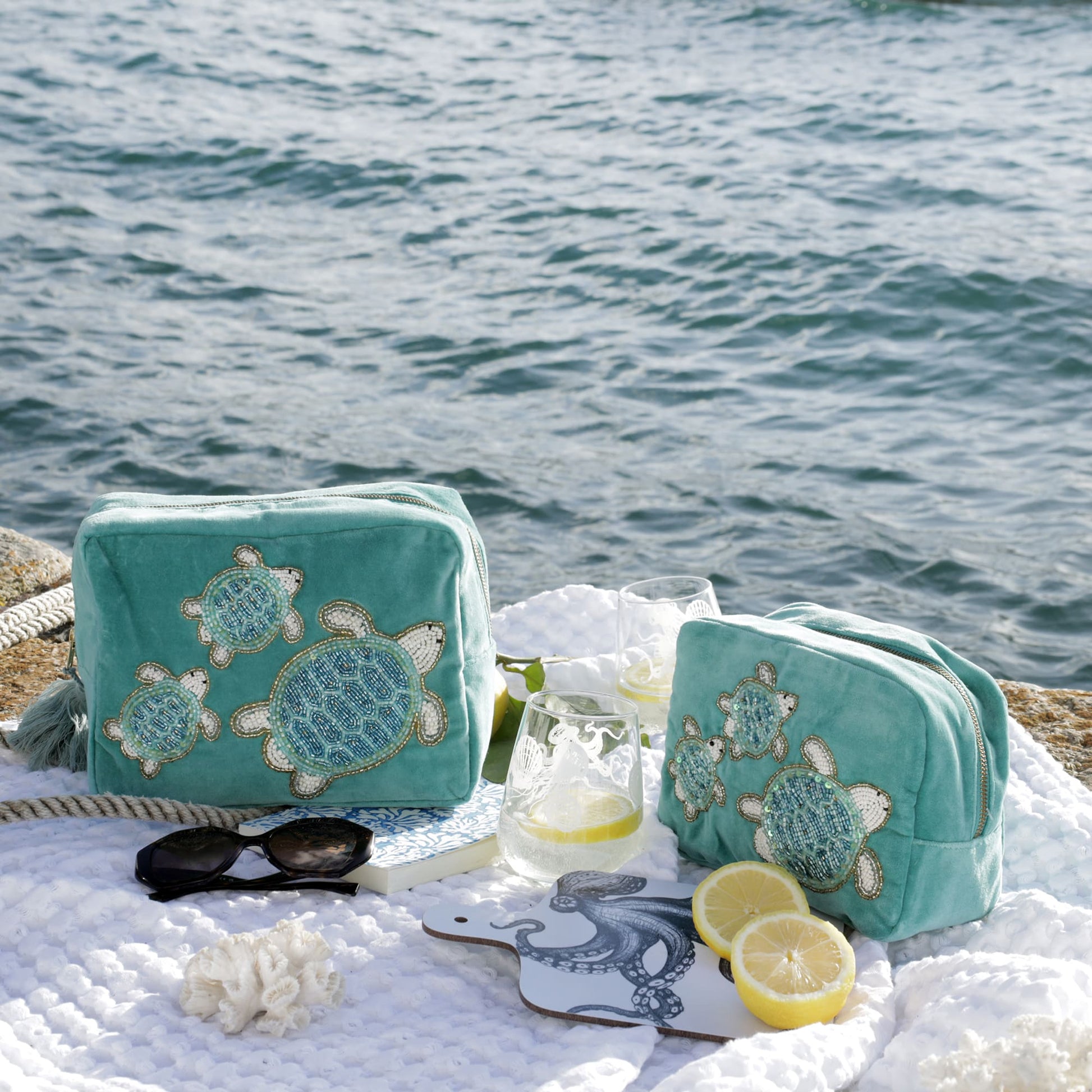 A scenic coastal flatlay featuring two turquoise velvet bags adorned with beaded sea turtle designs, set against a backdrop of the ocean. The bags rest on a soft white textured blanket alongside nautical accents, including a pair of black sunglasses, a small coral ornament, lemon halves, two etched drinking glasses with lemon slices, a rope, and a white mini chopping board illustrated with a blue octopus. The setup evokes a serene, beachy vibe perfect for summer travel or seaside relaxation.