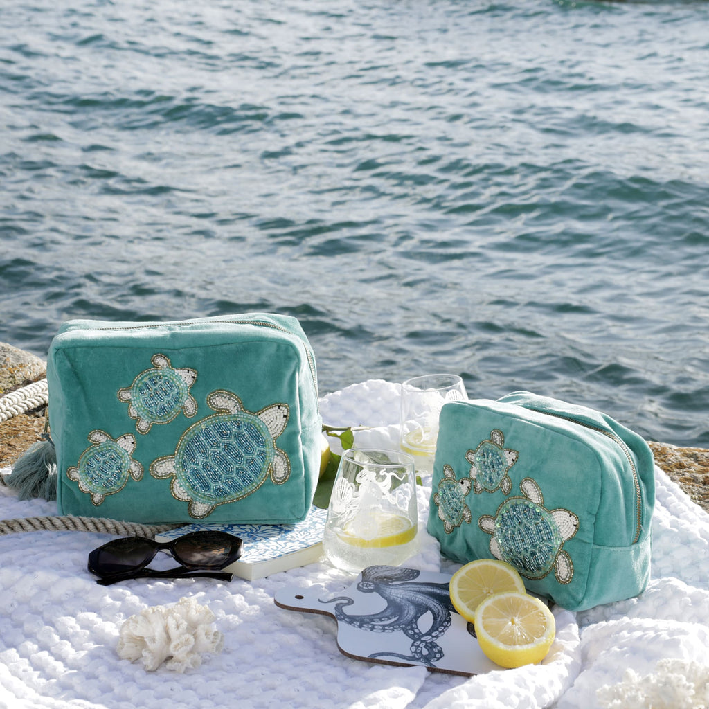 Two aqua green velvet bags featuring beaded sea turtle designs are displayed on a textured white blanket by the sea. The scene is styled with nautical rope, black sunglasses, a blue patterned notebook, lemon halves, a glass of lemon water, and a white cutting board with an illustrated blue octopus, all set against a sparkling ocean backdrop evoking a fresh, coastal summer atmosphere.