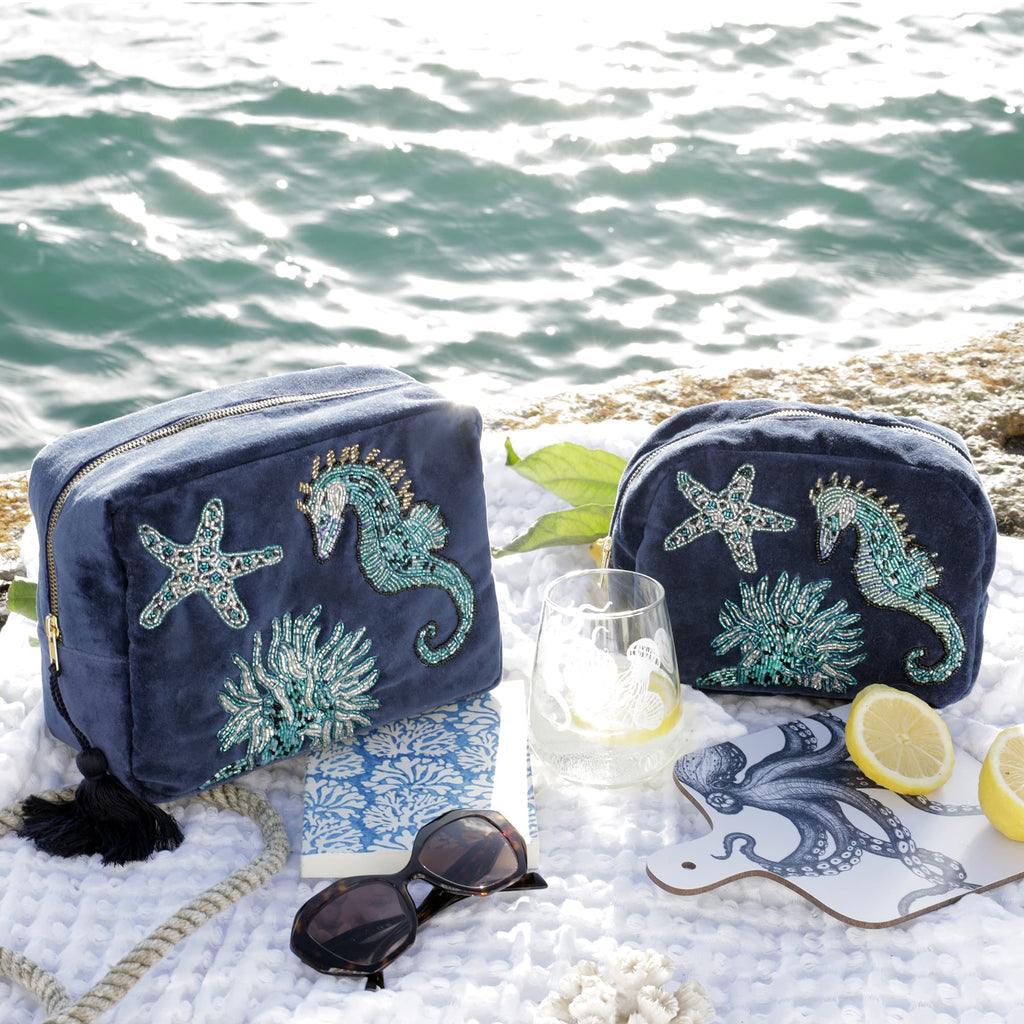 Two navy velvet bags decorated with shimmering beaded designs of seahorses, starfish, and coral sit on a white textured blanket beside the sea. The scene includes a nautical rope, black sunglasses, a blue coral-patterned notebook, a glass with a lemon slice, a white chopping board illustrated with a blue octopus, and lemon halves, all basking in natural sunlight with the ocean in the background. The composition evokes a relaxed, coastal summer vibe.