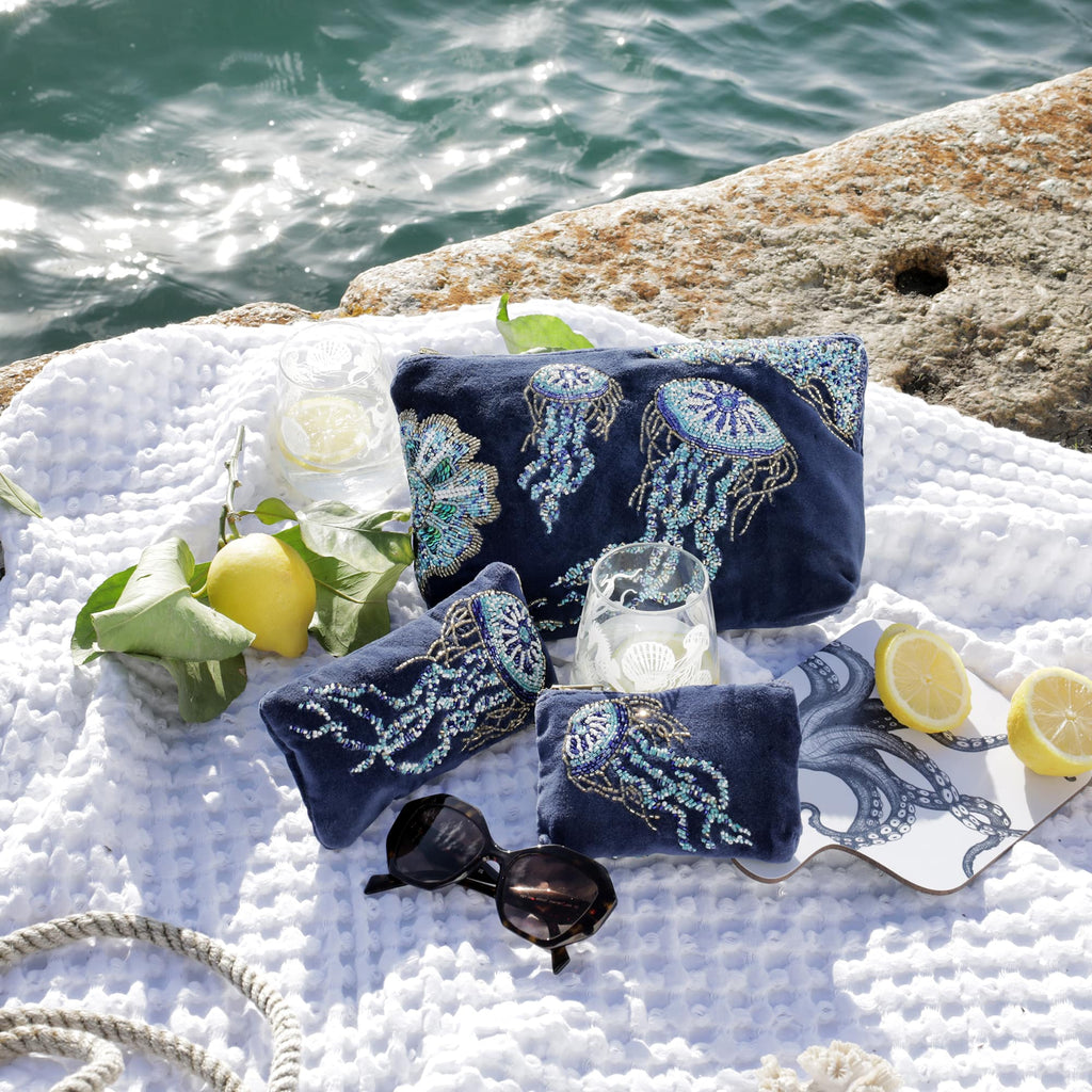 A seaside flatlay featuring a collection of navy velvet beaded accessories with intricate jellyfish designs, including a wash bag, purse, and glasses case. The items are styled on a white textured blanket with lemons, sunglasses, two etched glasses with drinks, and a coaster featuring an octopus illustration, all set against a sparkling turquoise sea and a stone ledge.