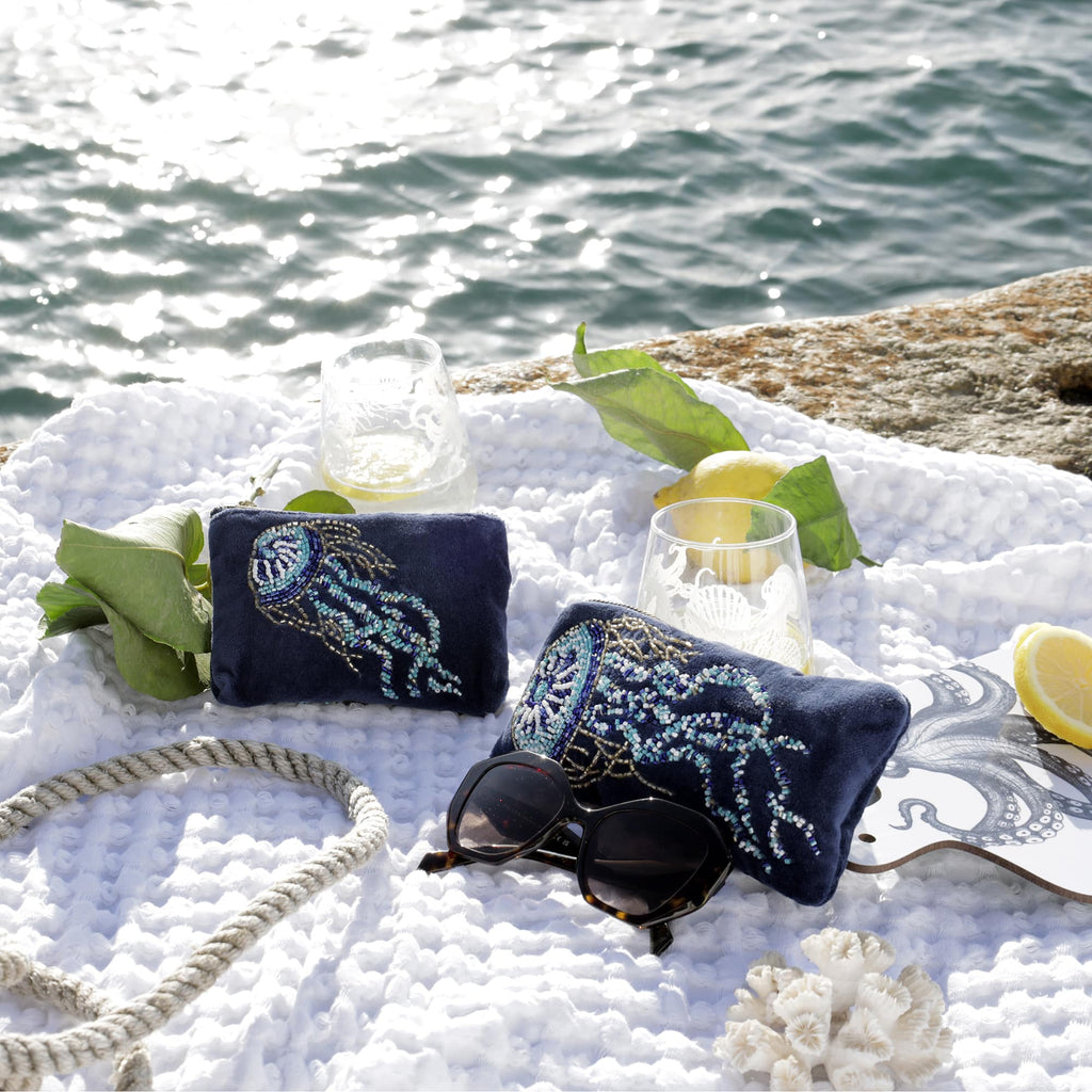 A close-up seaside flatlay showcasing two navy velvet purses with intricate beaded jellyfish designs, placed on a soft white textured blanket. The scene includes etched glasses with drinks, fresh lemons with leaves, sunglasses, a rope, a decorative octopus coaster, and coral, all set against a shimmering ocean backdrop and a rocky edge.