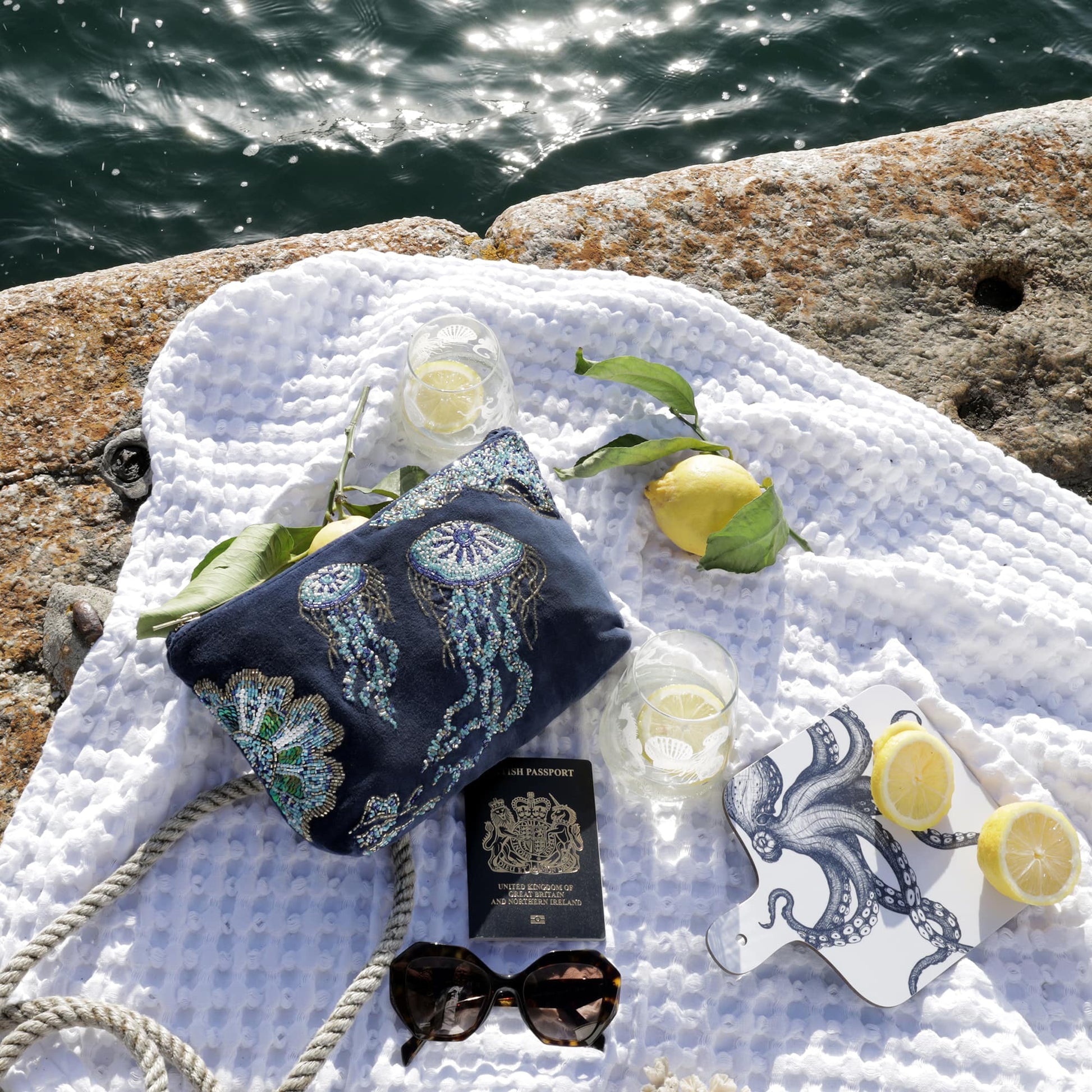 A coastal flatlay scene featuring a navy velvet bag adorned with beaded jellyfish and sea life designs, resting on a white textured blanket. Surrounding items include a British passport, dark sunglasses, two etched drinking glasses with lemon slices, fresh lemons with leaves, a decorative octopus coaster, and a thick nautical rope, all set beside a sunlit, rippling sea and a rugged stone edge.