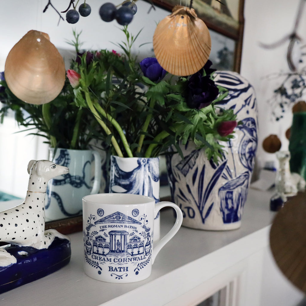 Bath Mug on a mantlepiece with an octopus jug containing flowers.Also on the mantlepiece is a statue of a spotted dog and a blue & white lidded Jar in front of a mirror. 