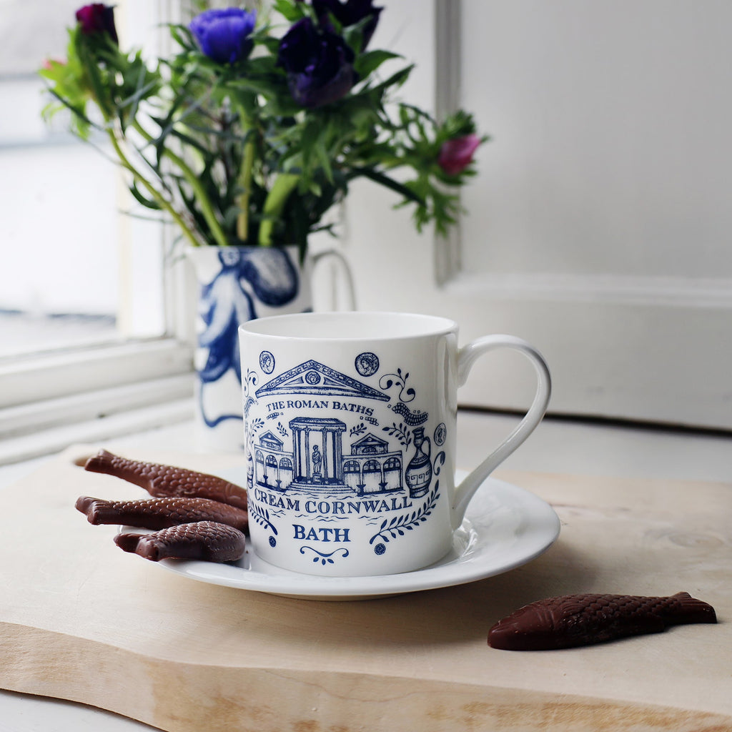 Bone China Bath Mug  placed on a white saucer with some chocolate shaped as fish.In the distance you can see an Octopus vase containing flowers placed on a table.