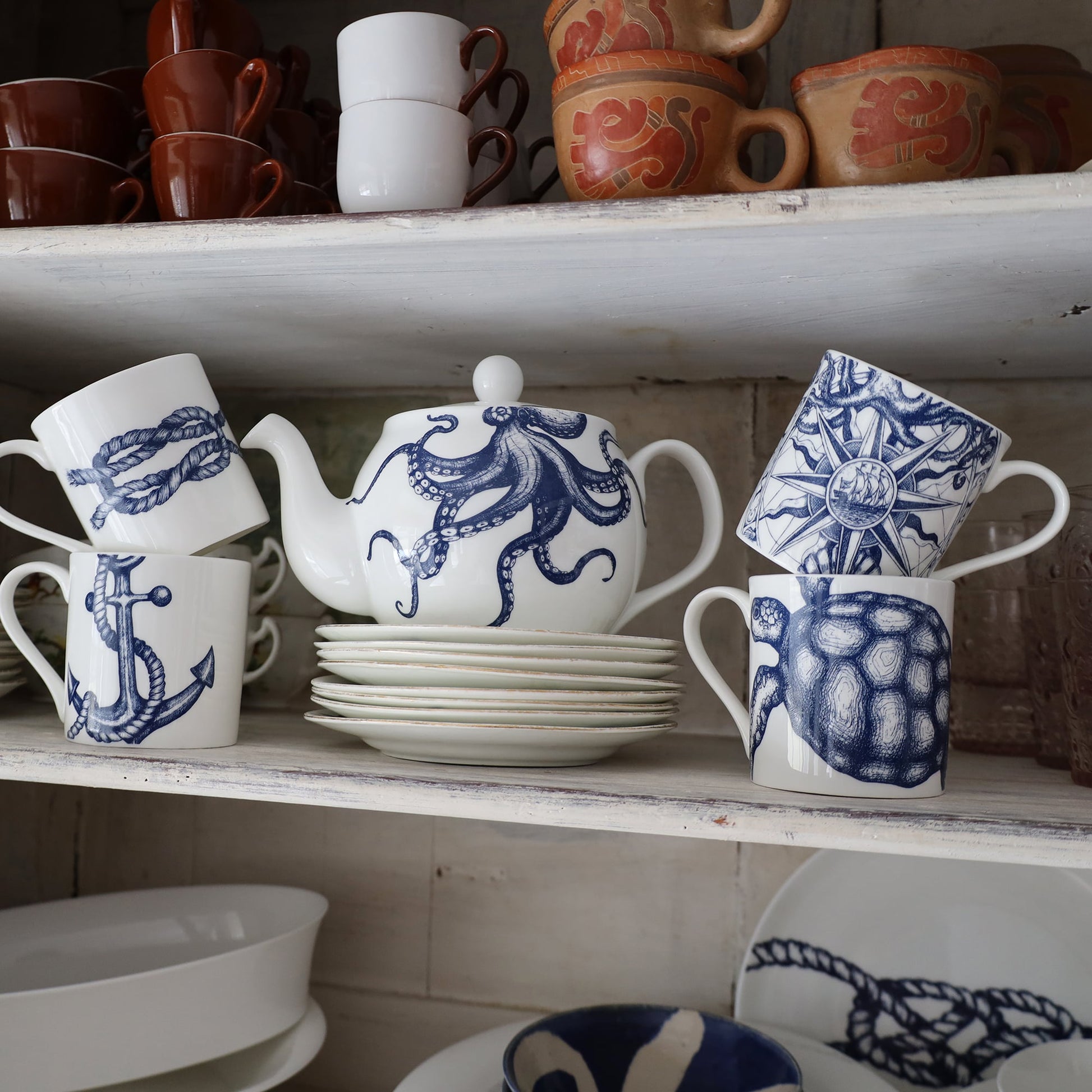 Turtle mug place next to an Octopus Teapot which is on a stack of small white plates. They are on a shelf,on the shelf above you can see espresso cups and other cups stacked.Below on another shelf  are other plates and bowls.