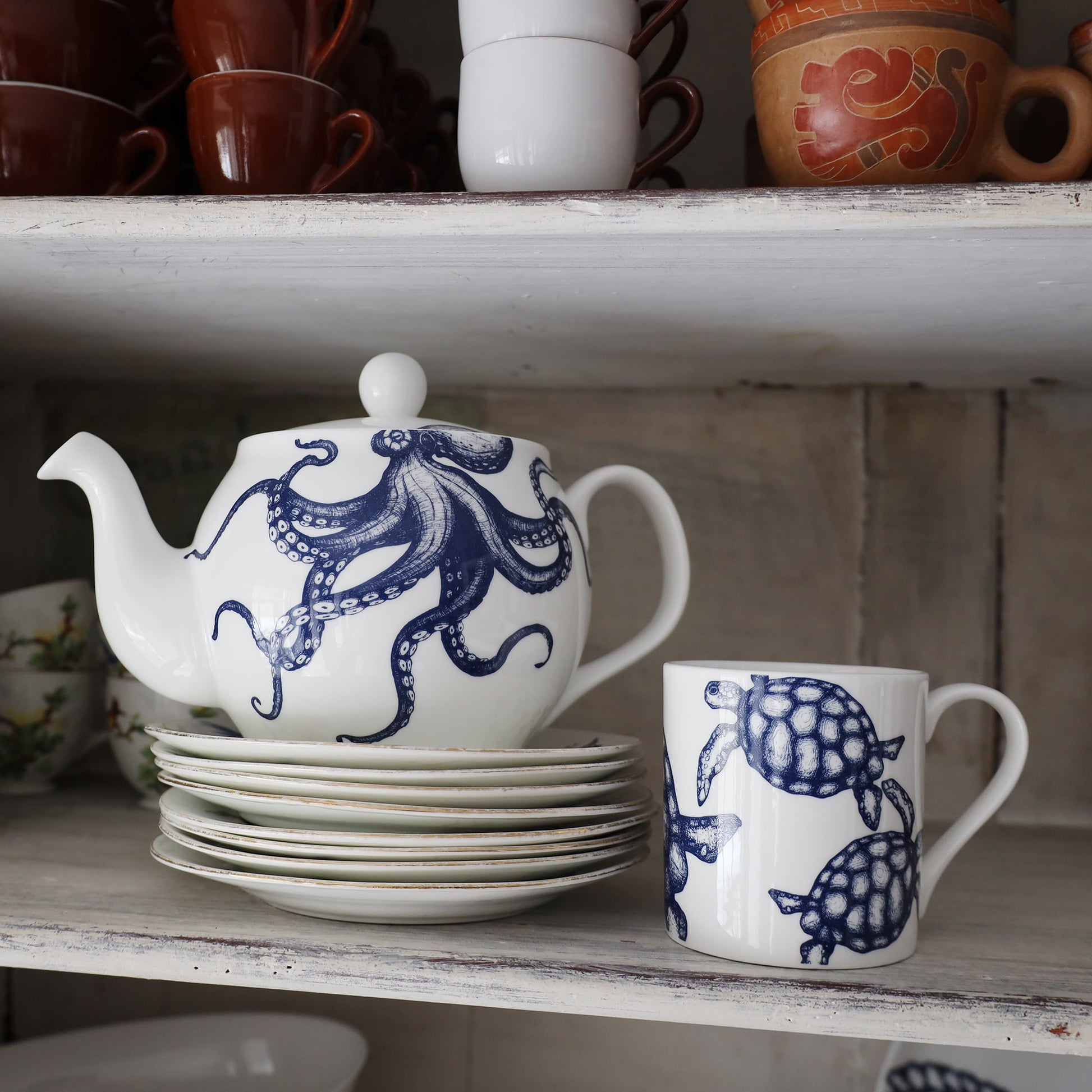 Turtle mug place next to an Octopus Teapot which is on a stack of small white plates. They are on a shelf,on the shelf above you can see espresso cups and other cups stacked.