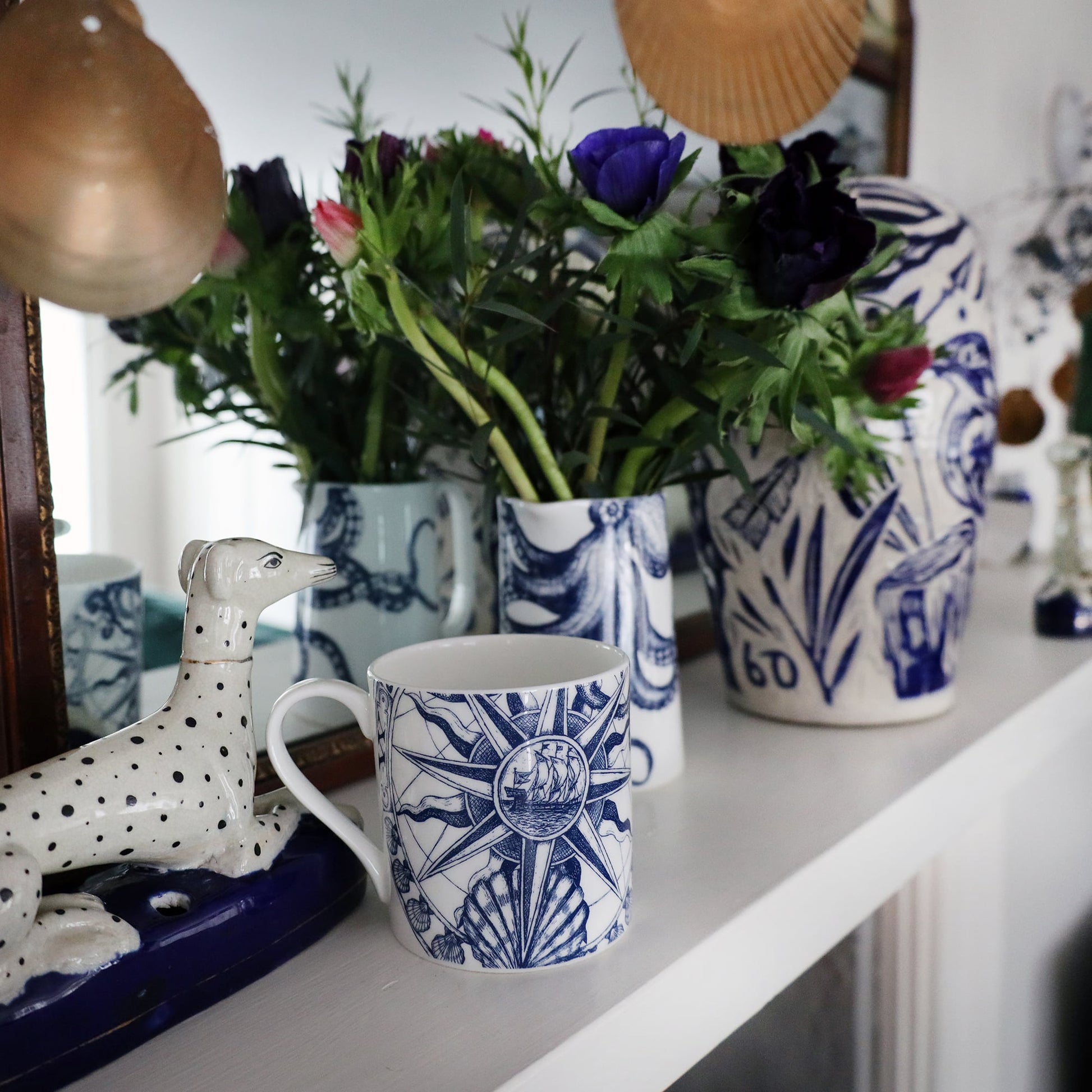 Compass  Mug on a mantlepiece with an octopus jug containing flowers.Also on the mantlepiece is a statue of a spotted dog and a blue & white lidded Jar in front of a mirror.