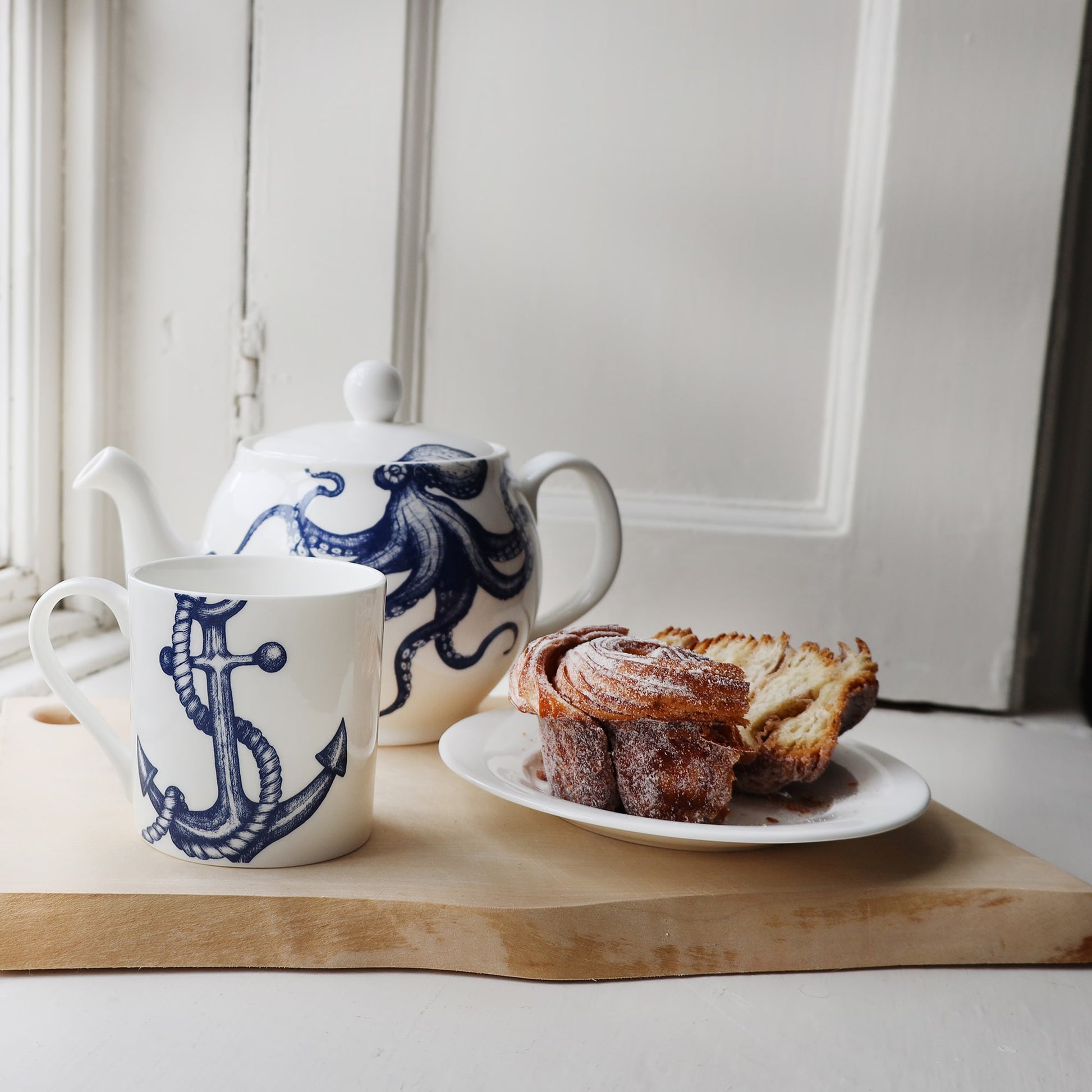 Anchor mug placed on a wooden board next to a plate with a split Cinnamon bun,also on the table is an Octopus Teapot .