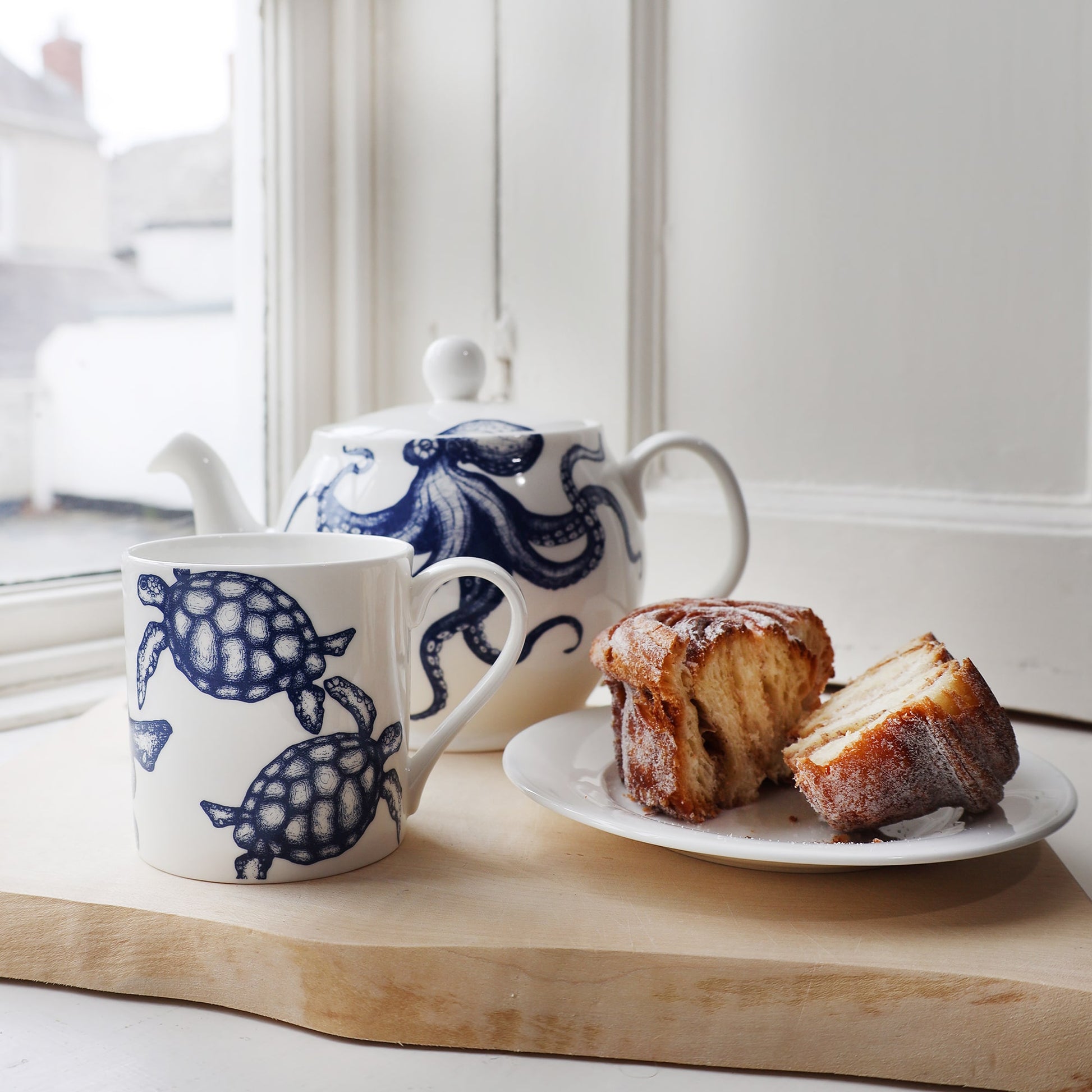 Turtle mug next to an Octopus Teapot placed on a wooden board next to a plate with a split  Cinnamon bun.