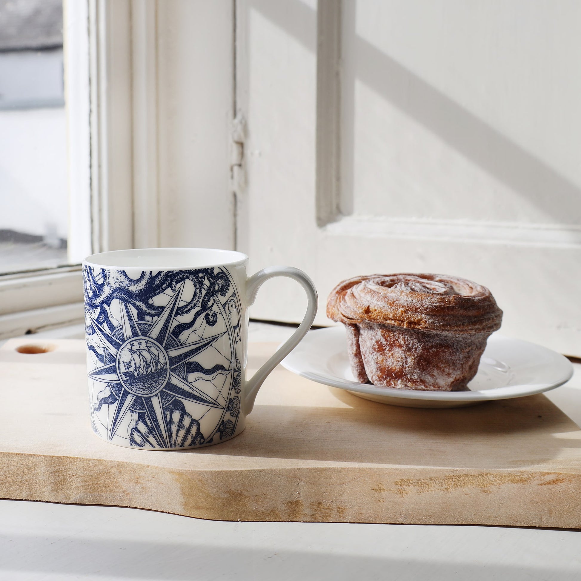 Compass mug  placed on a wooden board next to a plate with a Cinnamon bun.