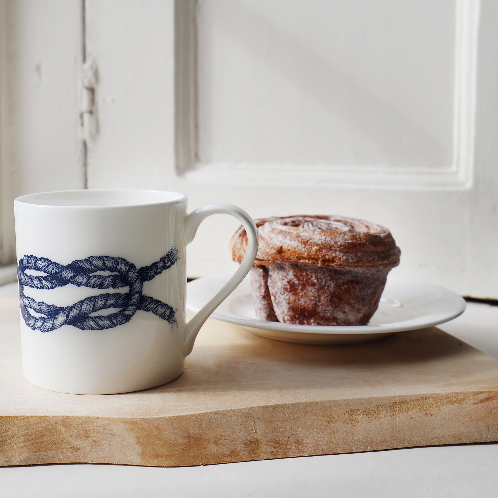 Knot mug  placed on a wooden board next to a plate with a Cinnamon bun.