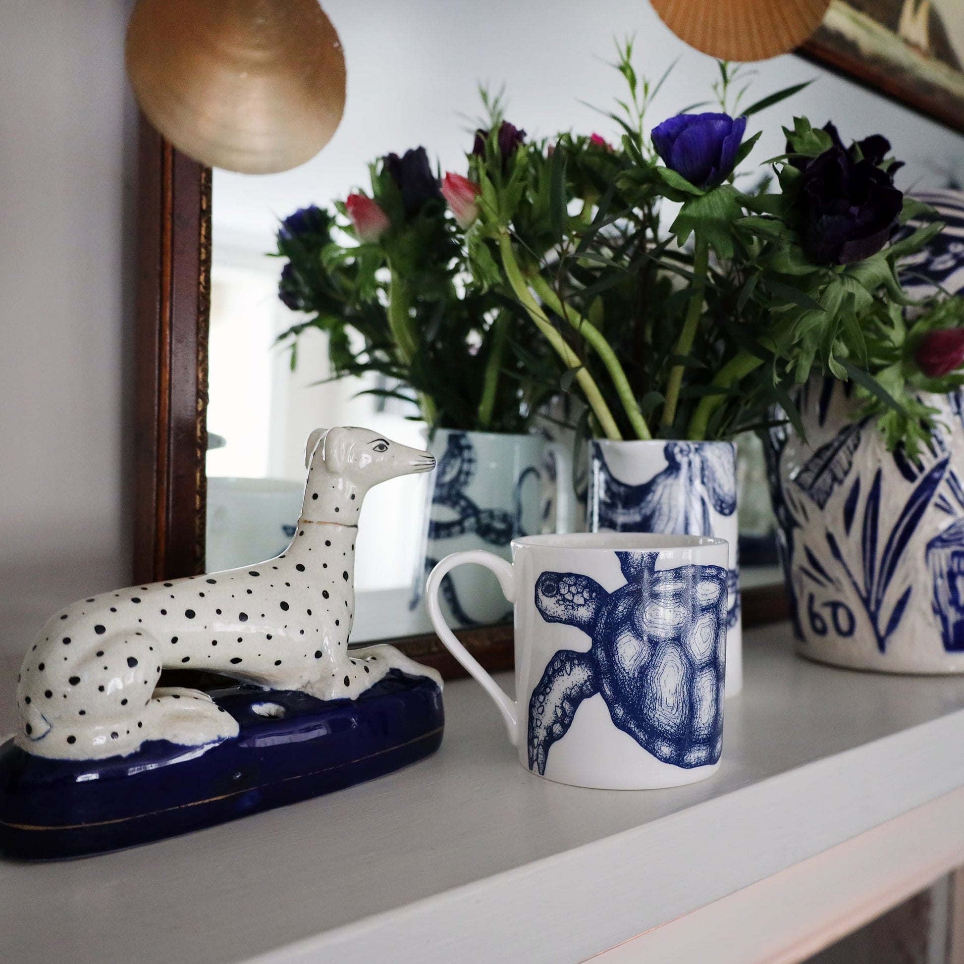 Turtle Mug on a mantlepiece with an octopus jug containing flowers.Also on the mantlepiece is a statue of a spotted dog and a blue & white lidded Jar in front of a mirror.