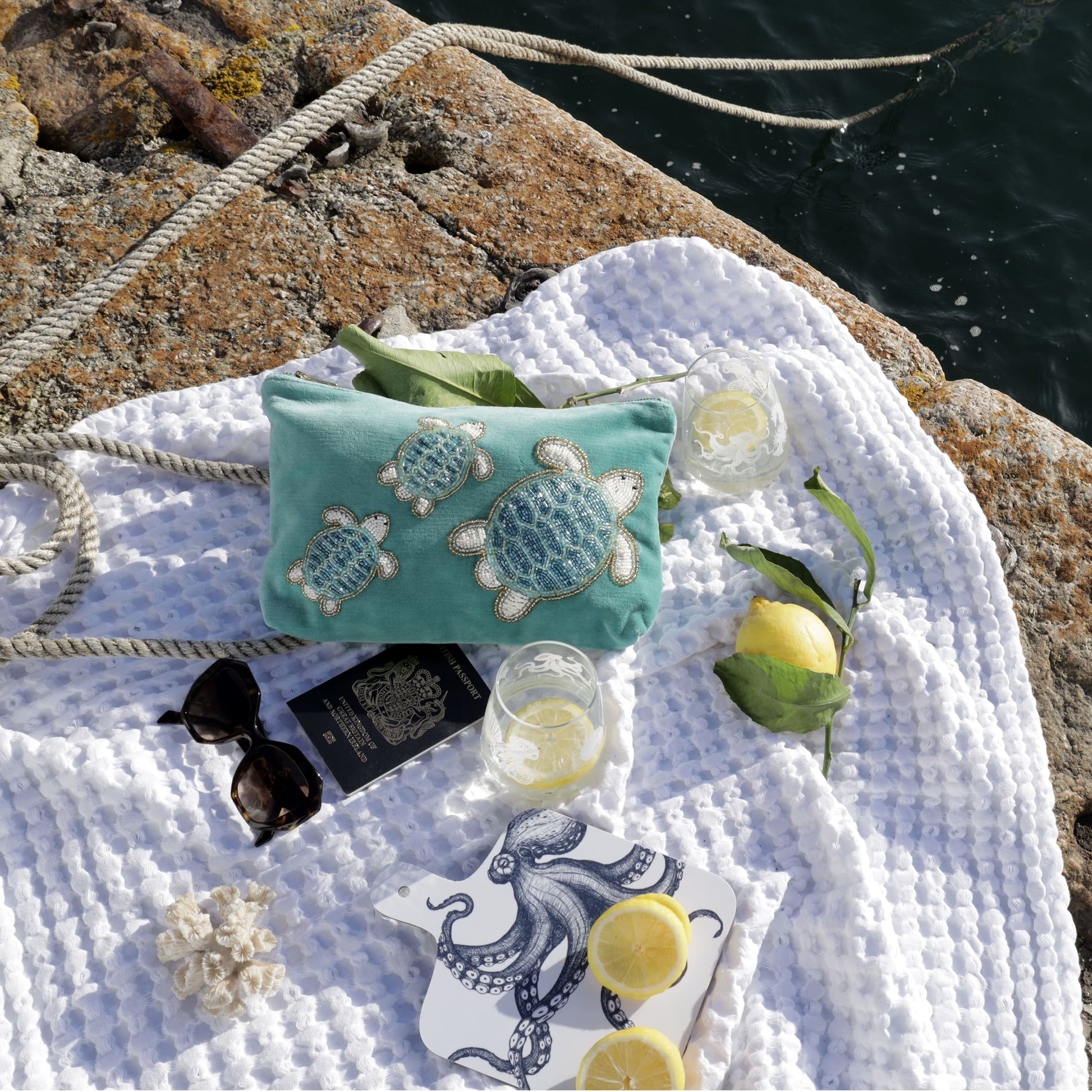 A seaside flatlay showcasing a turquoise velvet pouch adorned with three intricately beaded sea turtles, placed on a textured white blanket atop a rocky pier. Surrounding the pouch are nautical elements including coiled rope, a pair of sunglasses, a British passport, two etched glasses with lemon slices, a lemon with leaves, a small coral decoration, and a white cutting board featuring a navy octopus illustration with lemon halves on top. The sparkling ocean water glistens in the background.