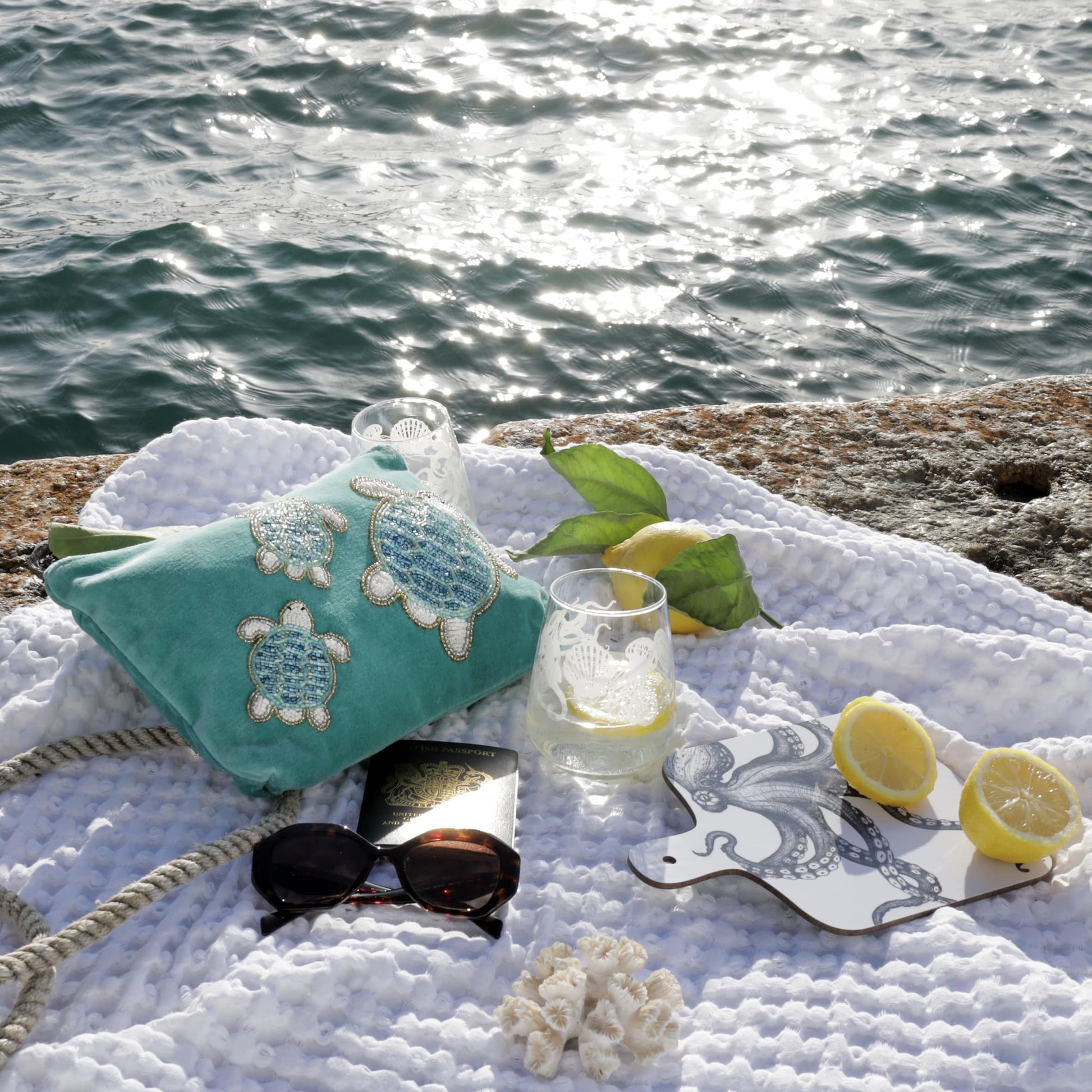 A scenic coastal flatlay featuring a turquoise velvet pouch embellished with three beaded sea turtles, resting on a white textured blanket beside the sea. Also pictured are a pair of sunglasses, a British passport, two etched glasses with lemon water, fresh lemons with leaves, a white and navy octopus-themed coaster, a length of rope, and small pieces of coral, all bathed in warm sunlight with the sea glistening in the background.