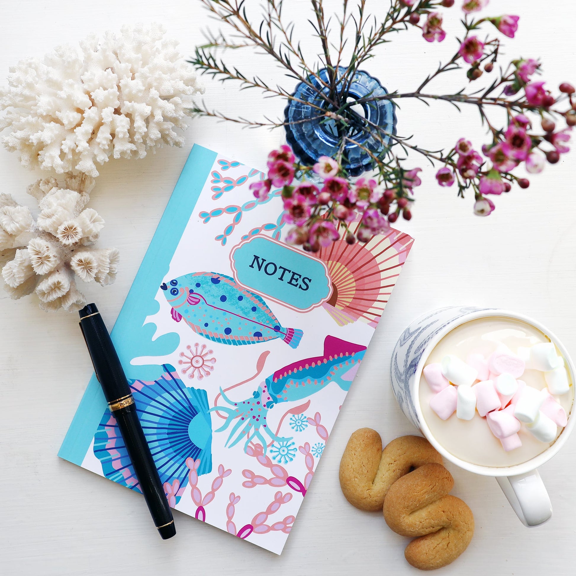 Overhead view of a flat lay featuring a vibrant "Catch of the Day" notebook with colorful illustrations of a flat fish, squid, scallop shells, and coral. The notebook is paired with a black and gold fountain pen, a blue glass vase of pink flowers, white coral decor, a cup of hot chocolate topped with marshmallows, and two butter biscuits, all arranged on a white surface.