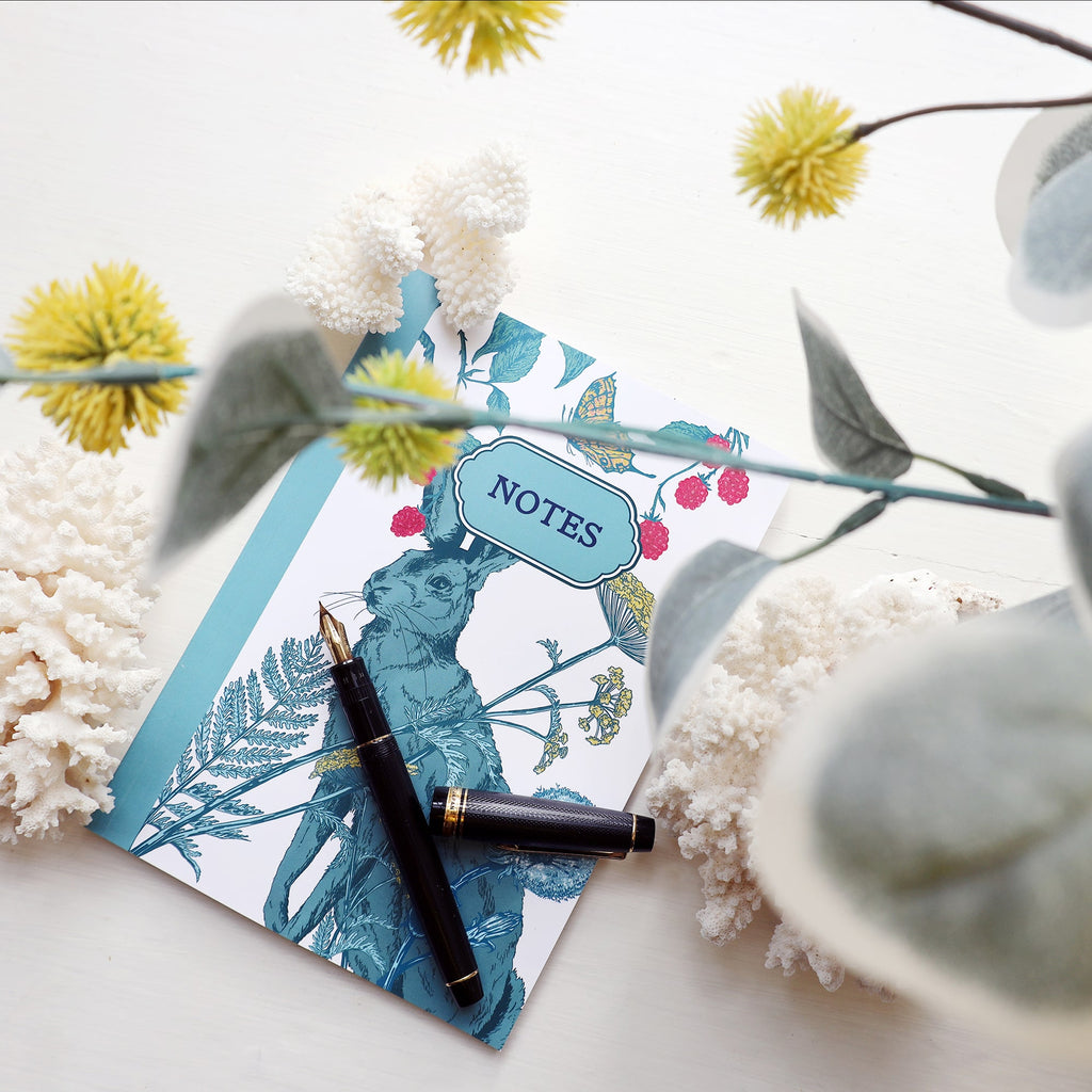 Hedgerow-themed notebook with an illustration of a teal hare, wildflowers, raspberries, and butterflies, placed on a white surface with coral pieces, a black fountain pen, and soft yellow blooms in the foreground.