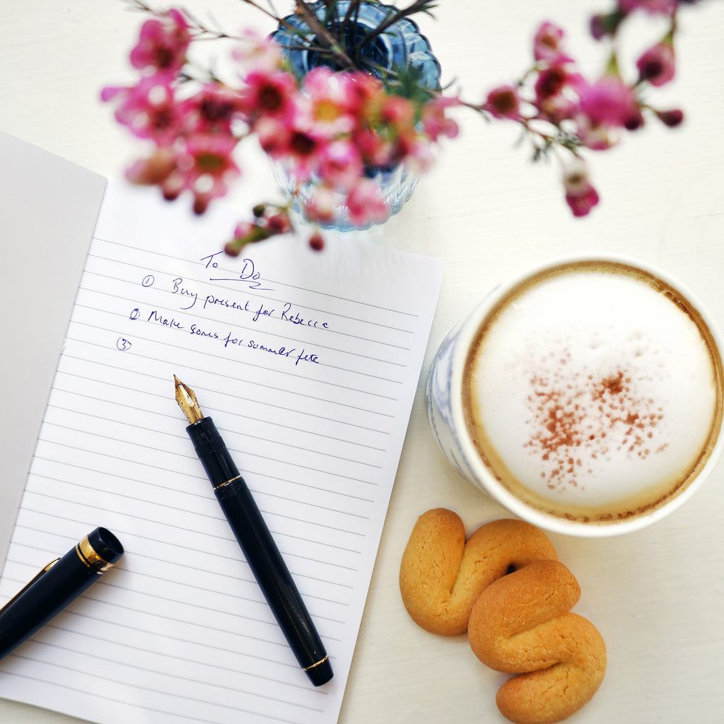 Open notebook with a handwritten to-do list, a fountain pen resting on the page, and a cappuccino with cinnamon beside two butter biscuits. Pink flowers in a small blue vase add a soft decorative touch to the scene.