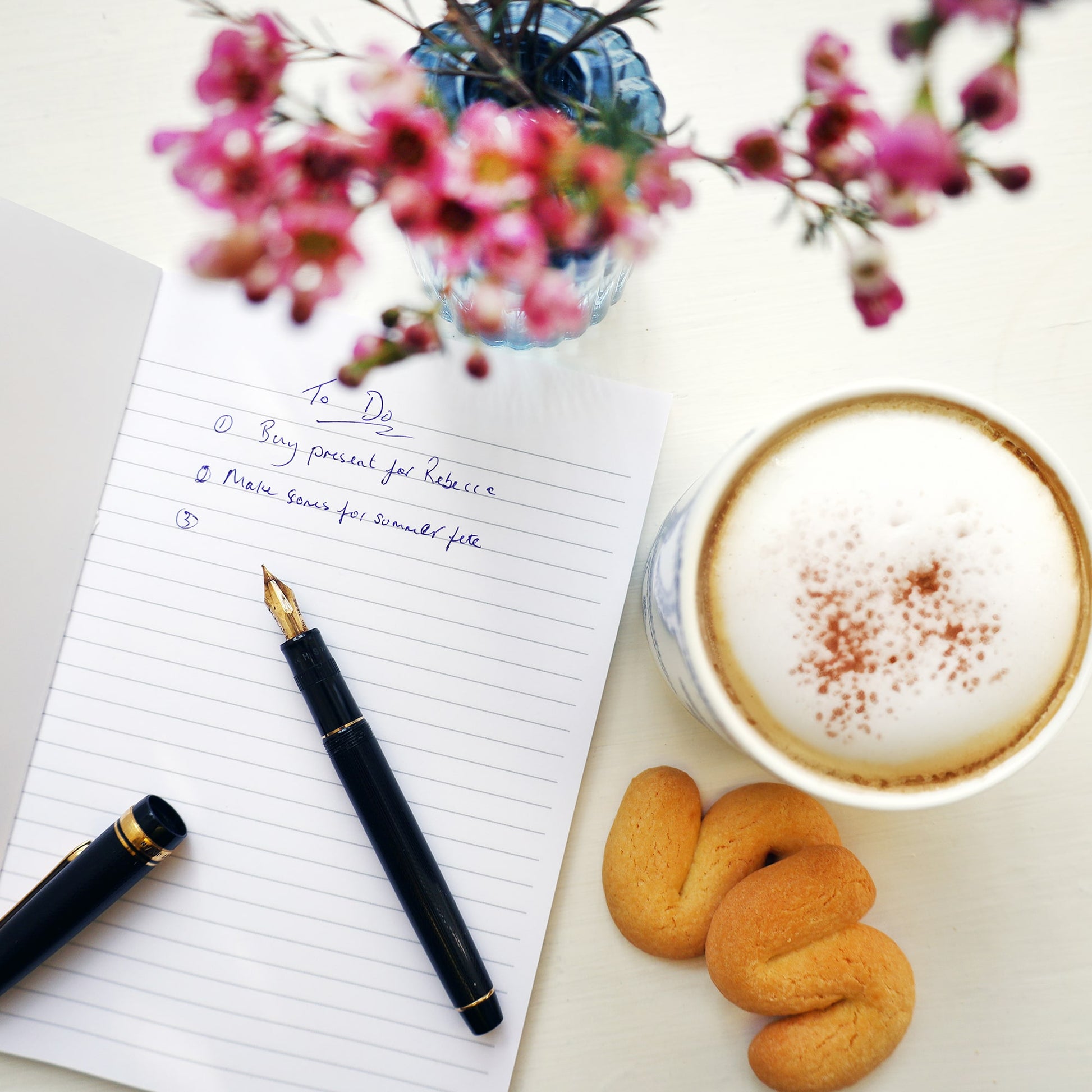  Open notebook with a handwritten to-do list, a fountain pen resting on the page, and a cappuccino with cinnamon beside two butter biscuits. Pink flowers in a small blue vase add a soft decorative touch to the scene.
