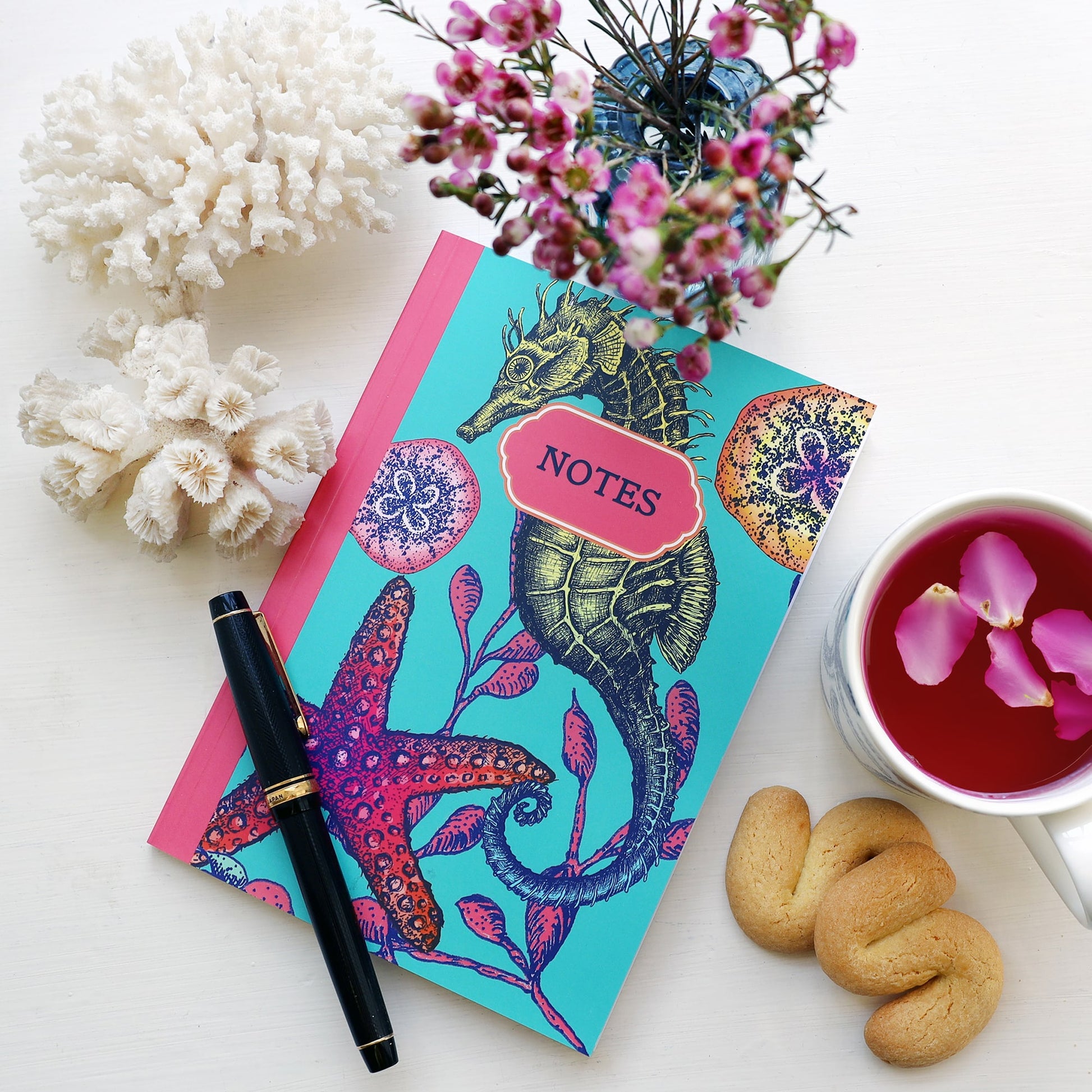 Flat lay of a vibrant sea-themed notebook with a turquoise cover featuring a seahorse, red starfish, and coral illustrations, labeled "NOTES" in a coral pink frame. Surrounding the notebook are a black fountain pen, white coral pieces, a blue vase with pink flowers, a cup of red herbal tea with rose petals, and two golden-brown biscuits.