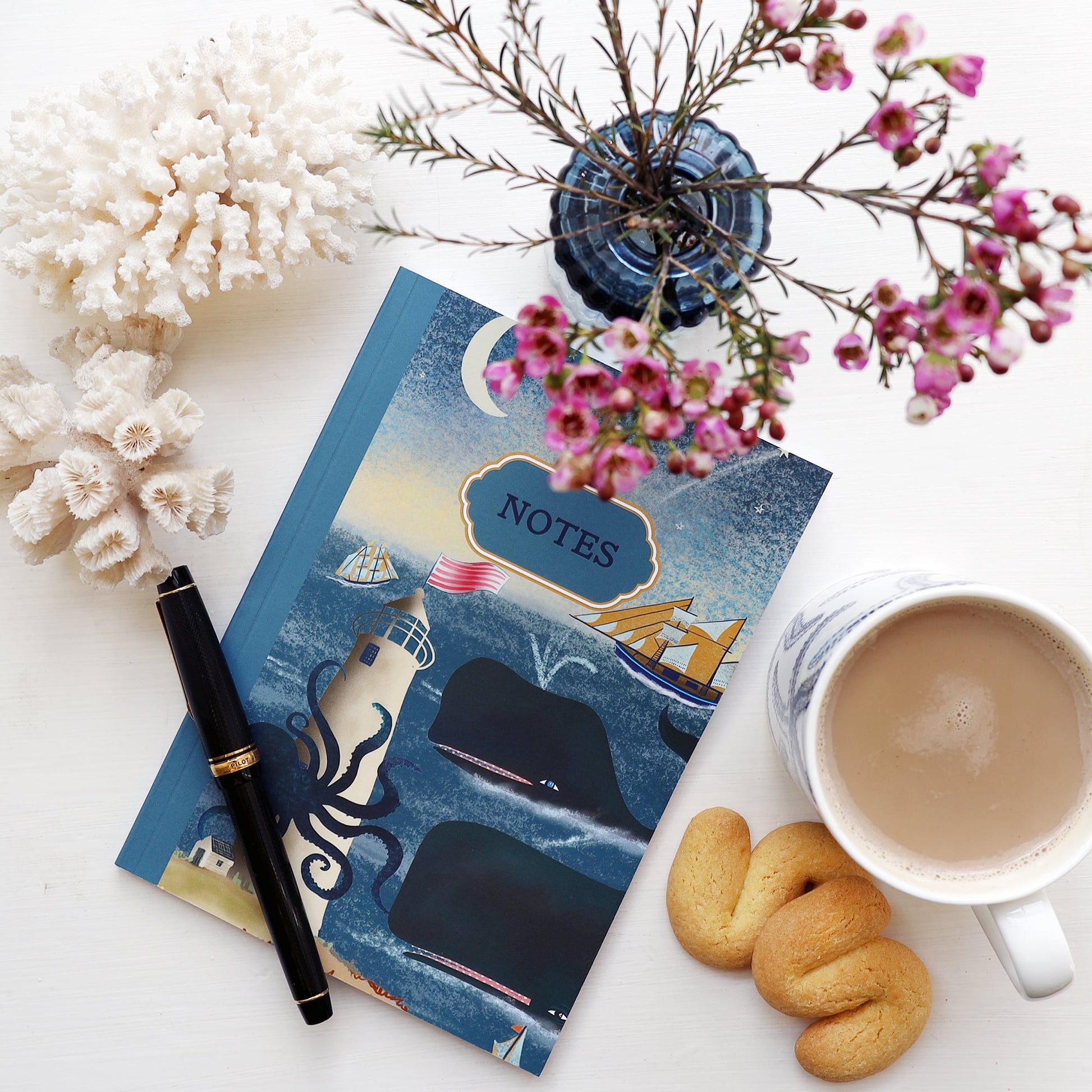 Flat lay of a whimsical ocean-themed notebook with an illustrated cover showing whales, a lighthouse, a ship, and an octopus, labeled "NOTES." The notebook is surrounded by a fountain pen, coral, a cup of coffee, flower vase, and a couple of butter biscuits on a white surface.
