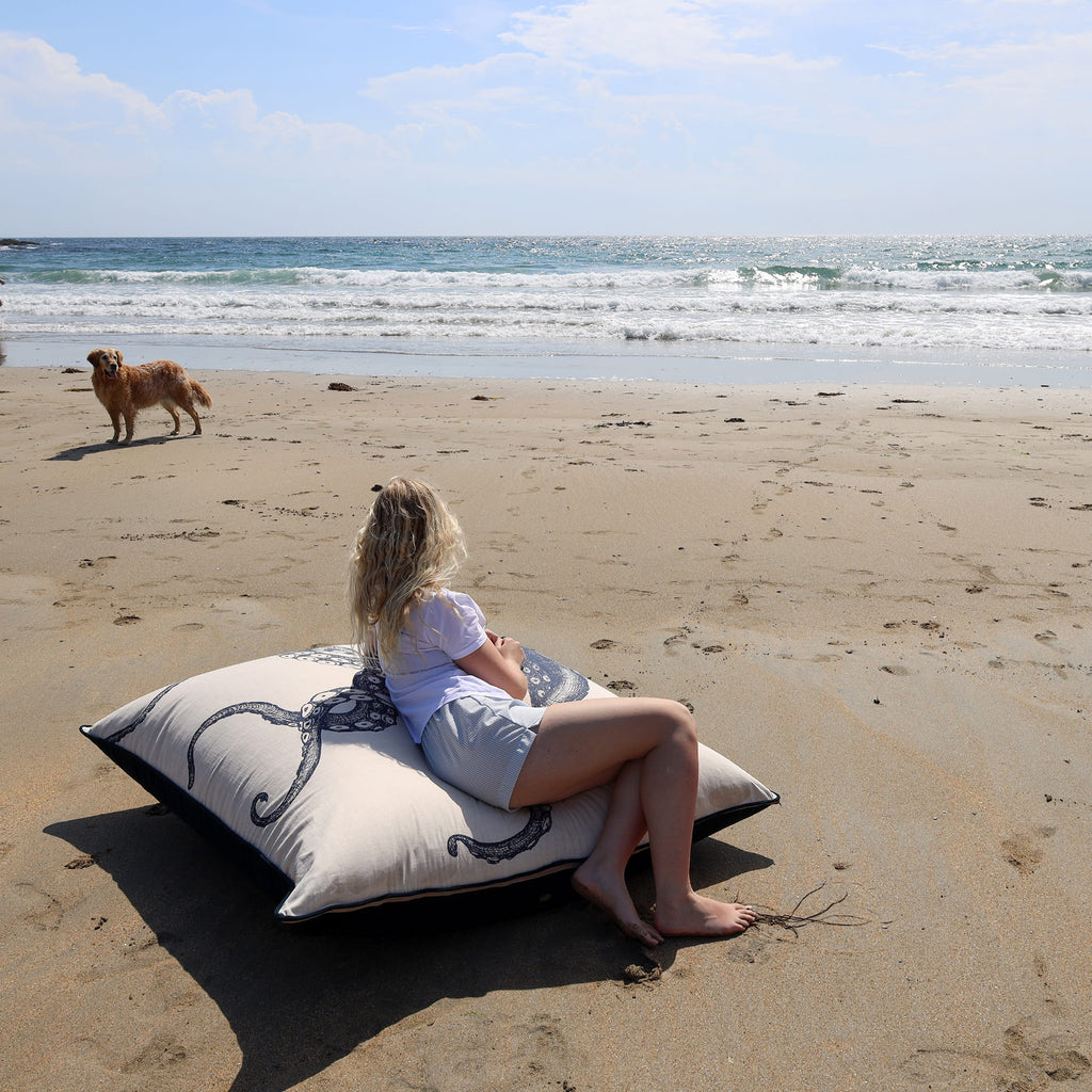 Woman sitting on a pillow on a sandy beach with a dog walking in the background