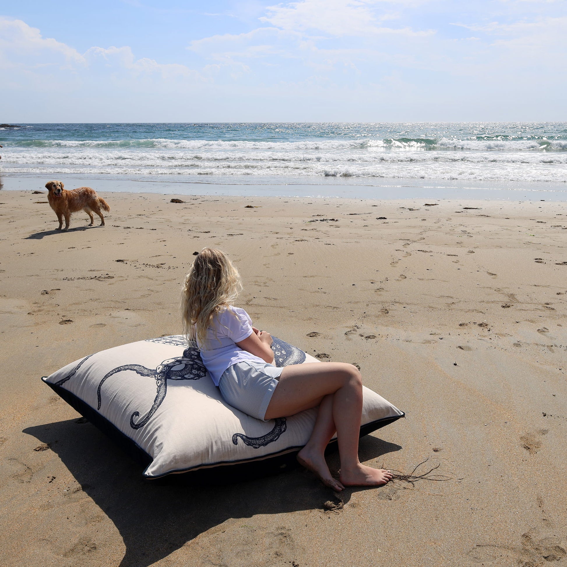 Woman sitting on a pillow on a sandy beach with a dog walking in the background