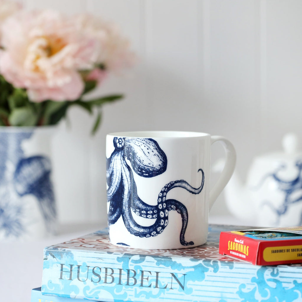 Close up of a white bone china mug with a blue illustrated octopus on it, sitting on a book with chocolate sardines and out of focus teapot & flowers in the background. 