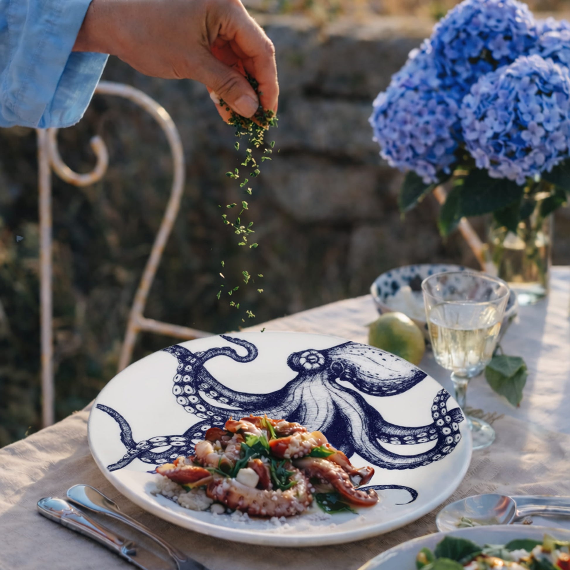 Person garnishing a dish on an octopus-themed plate with blue flowers and outdoor setting
