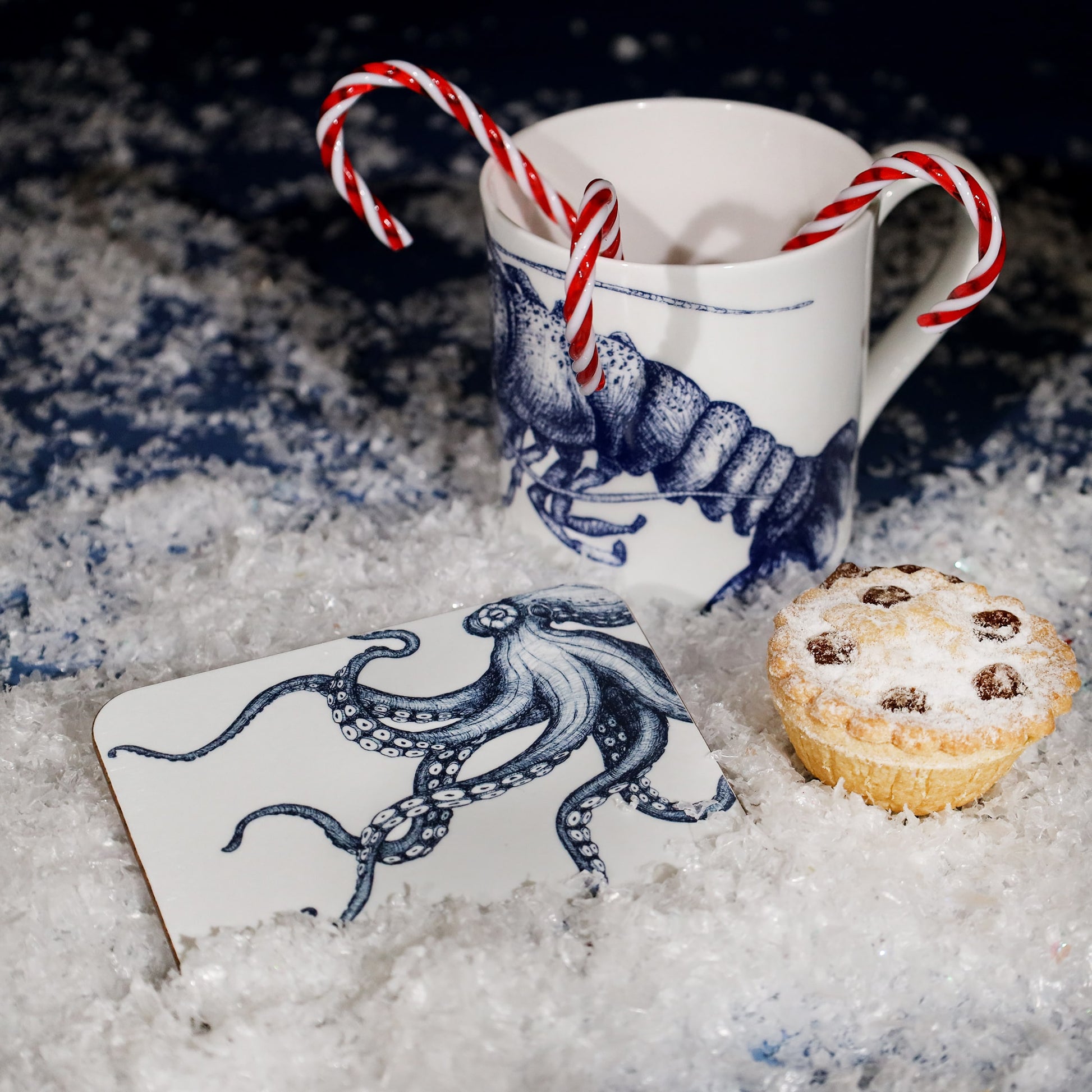 Festive scene featuring a white mug with a blue lobster illustration filled with candy canes, a matching coaster with an octopus design, and a mince pie dusted with sugar, all set on faux snow.