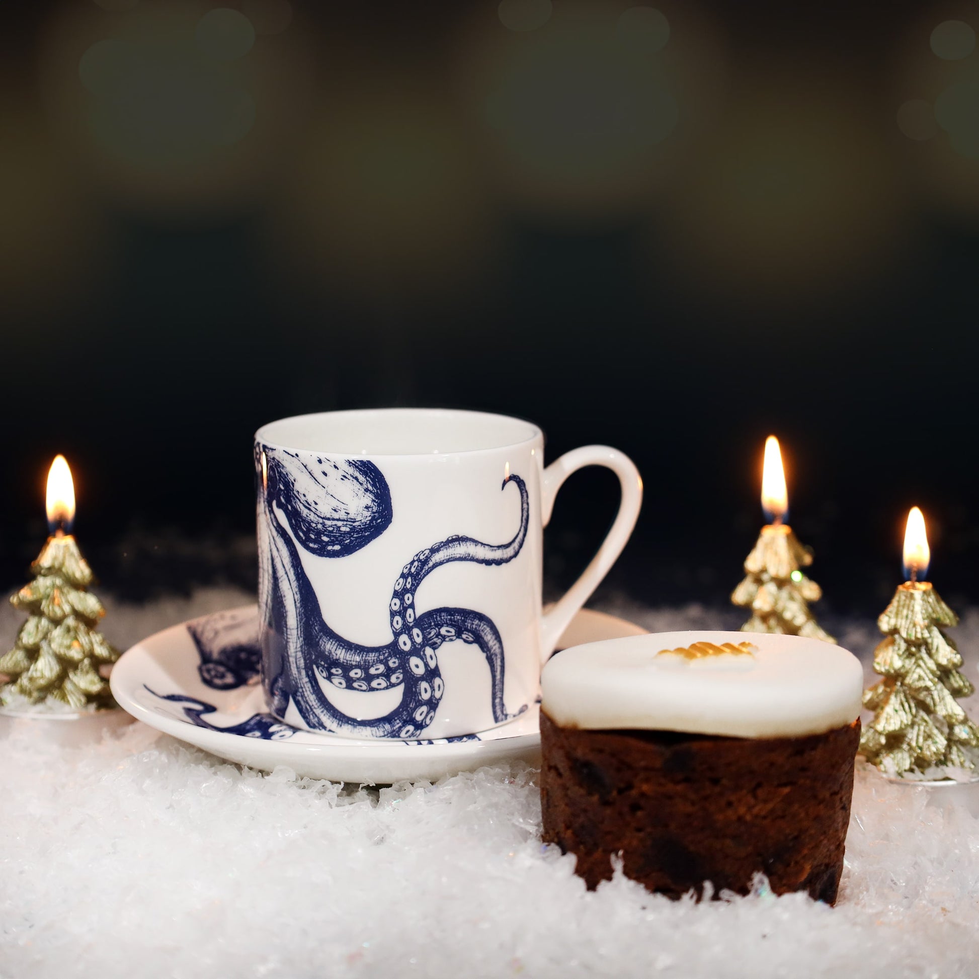 Festive scene featuring a white espresso cup and saucer with a blue octopus illustration, set on faux snow beside a small iced Christmas cake and surrounded by three lit gold tree-shaped candles.