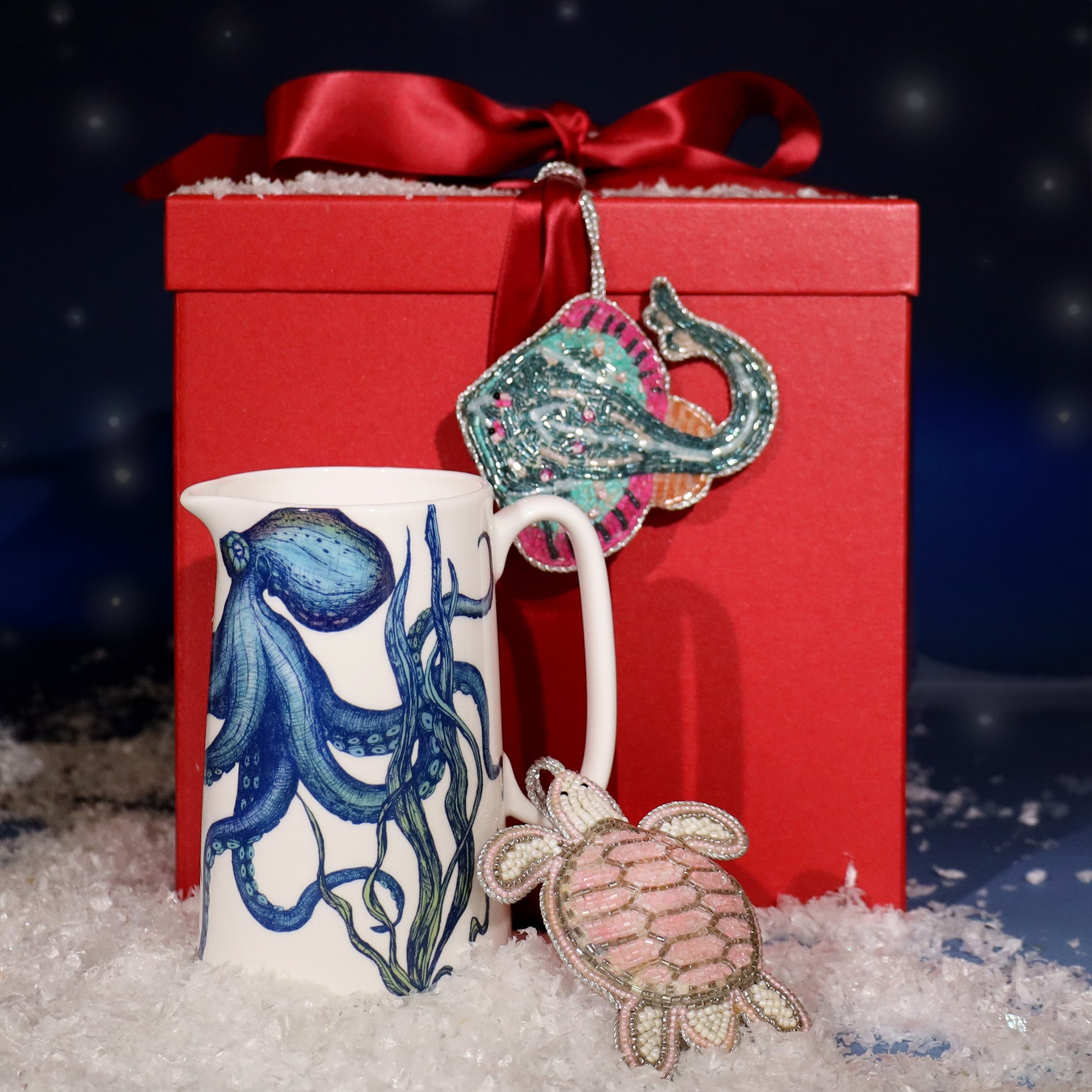 Festive display featuring a white jug with a blue octopus design, placed on faux snow in front of a red gift box tied with a satin ribbon, decorated with beaded sea-themed ornaments shaped like a turtle and a stingray.