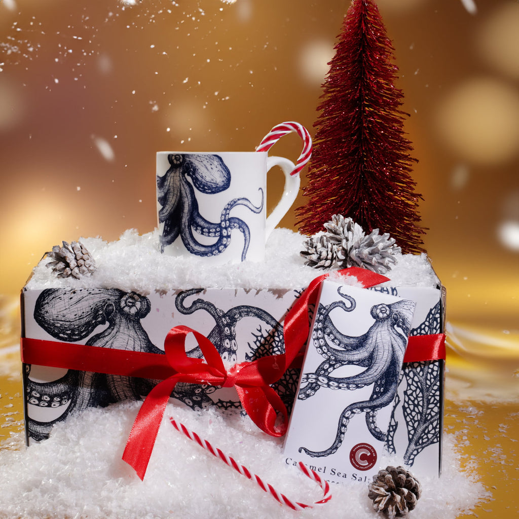 Octopus mug and chocolate gift set displayed in artificial snow with red ribbon, candy canes, pinecones, and a red Christmas tree, set against a golden festive background with softly falling snow and warm glowing lights.