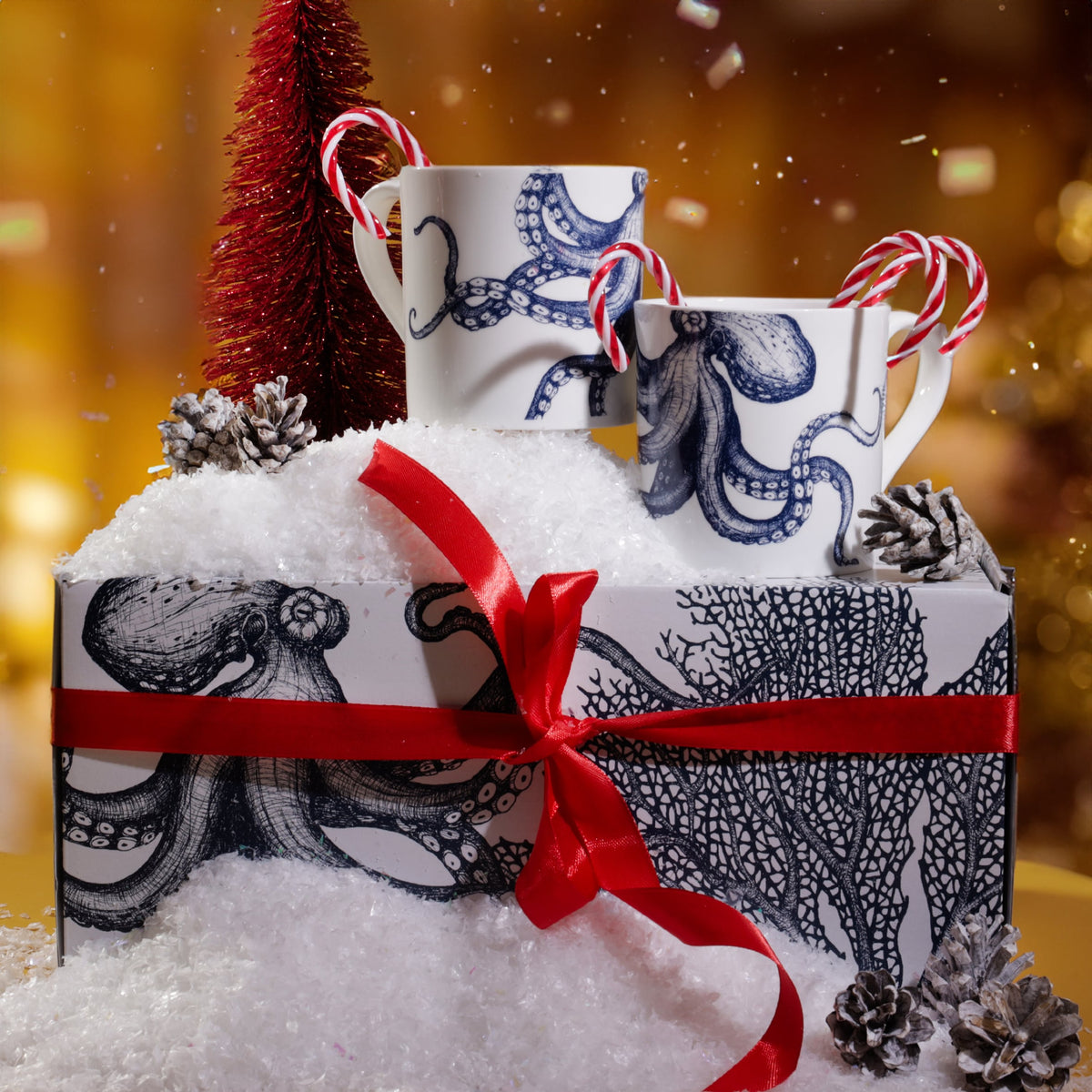 Octopus mug gift set displayed in a festive scene with artificial snow, red ribbon, candy canes, and a small red Christmas tree, set against a warm golden background with twinkling lights.