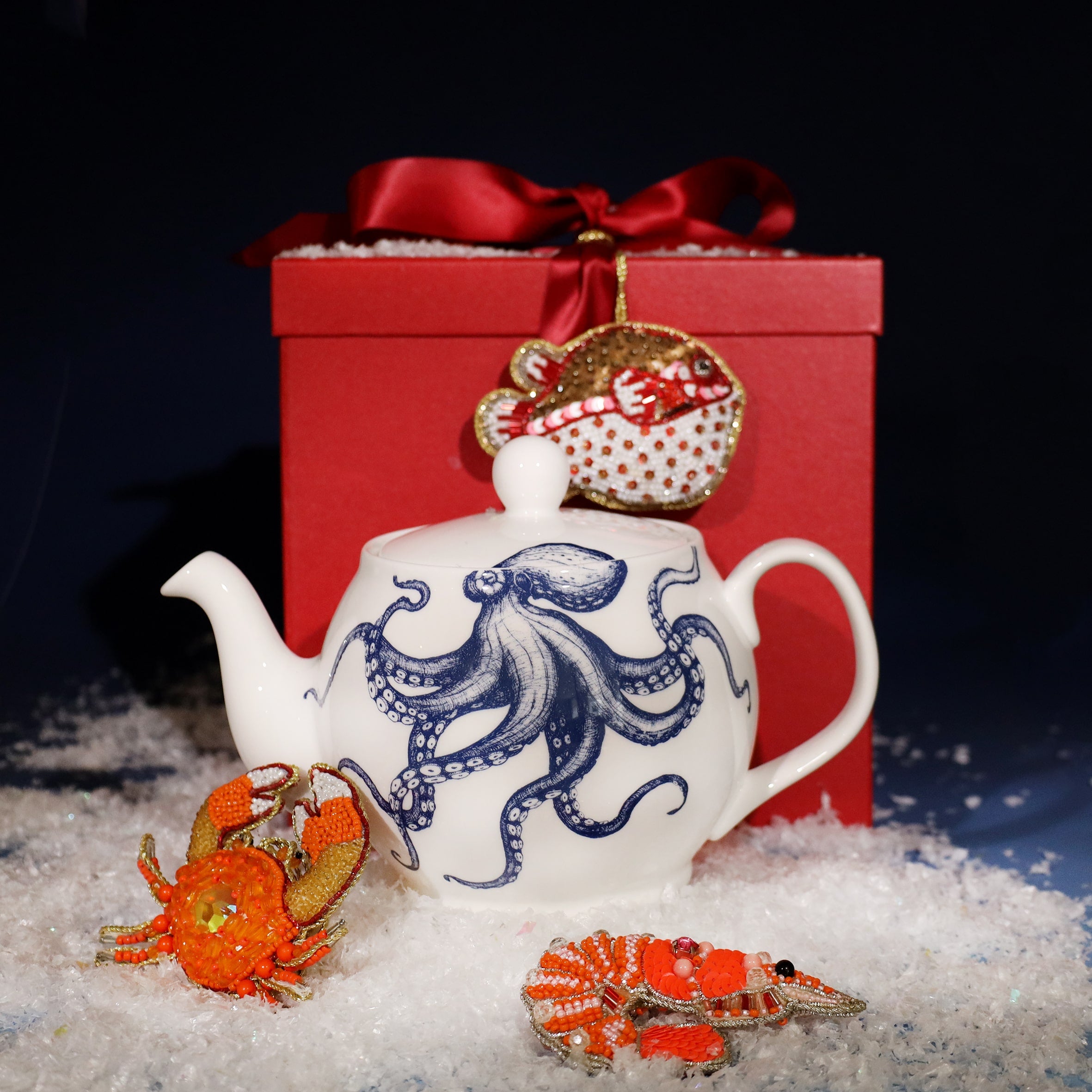 Festive scene featuring a white teapot with a blue octopus illustration, placed on faux snow in front of a red gift box tied with a satin ribbon, decorated with beaded sea creature ornaments including a crab, lobster, and fish.