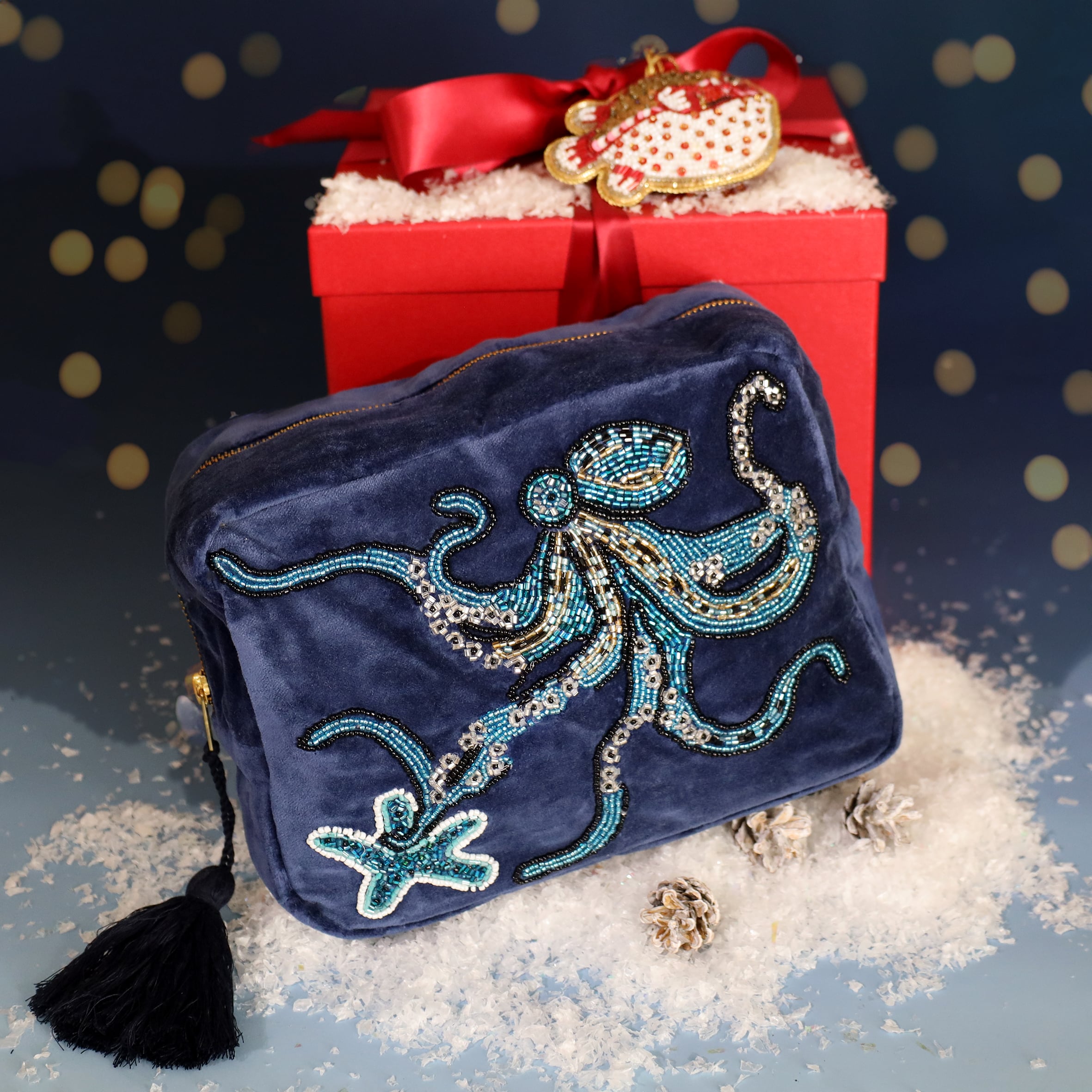 Festive display featuring a navy velvet wash bag embellished with a beaded octopus and starfish design, placed on faux snow in front of a red gift box tied with a satin ribbon and decorated with a beaded fish ornament.