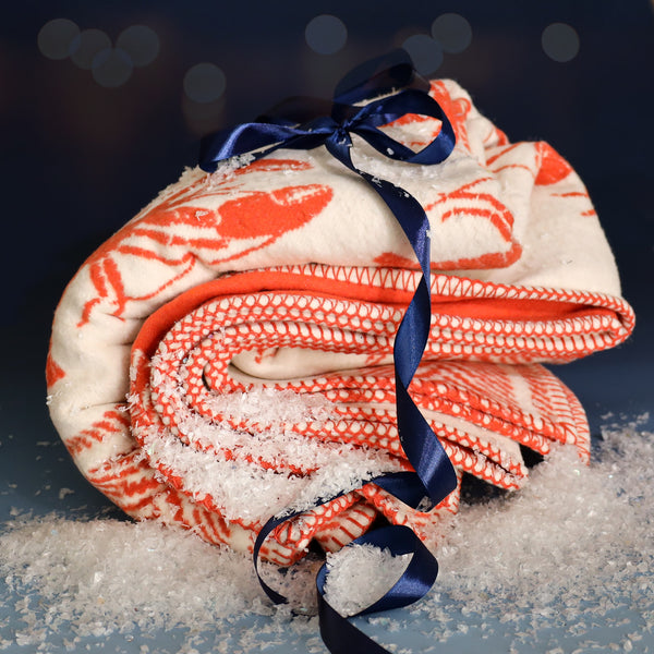 Folded cream and orange lobster-patterned throw tied with a navy blue ribbon, surrounded by faux snow against a softly lit festive background.
