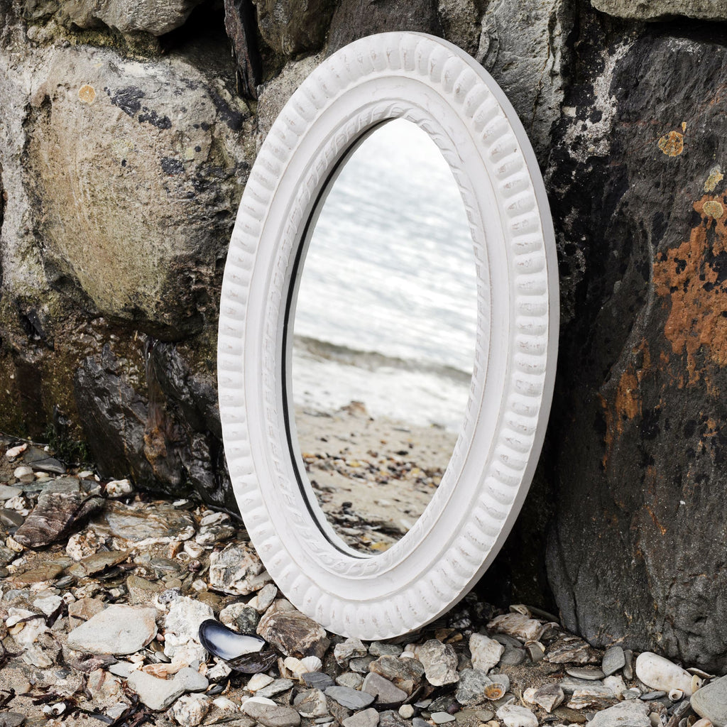 Oval whitewashed wooden mirror with rope detail carving propped up against the harbour wall on the beach with the shoreline reflected in it.