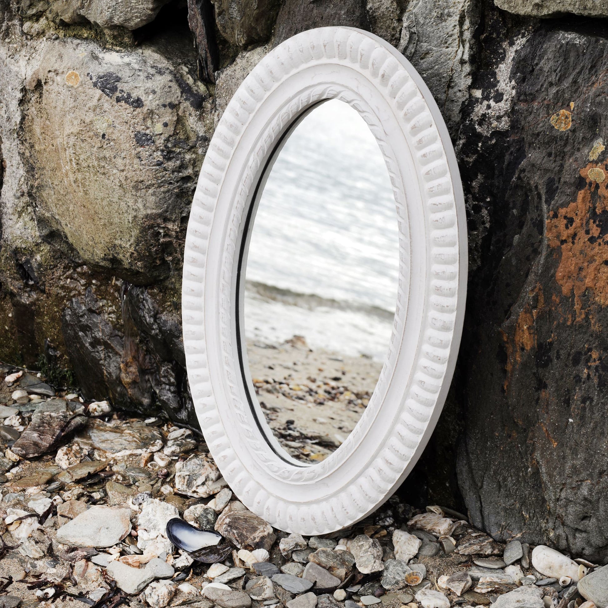 Oval whitewashed wooden mirror with rope detail carving propped up against the harbour wall on the beach with the shoreline reflected in it.