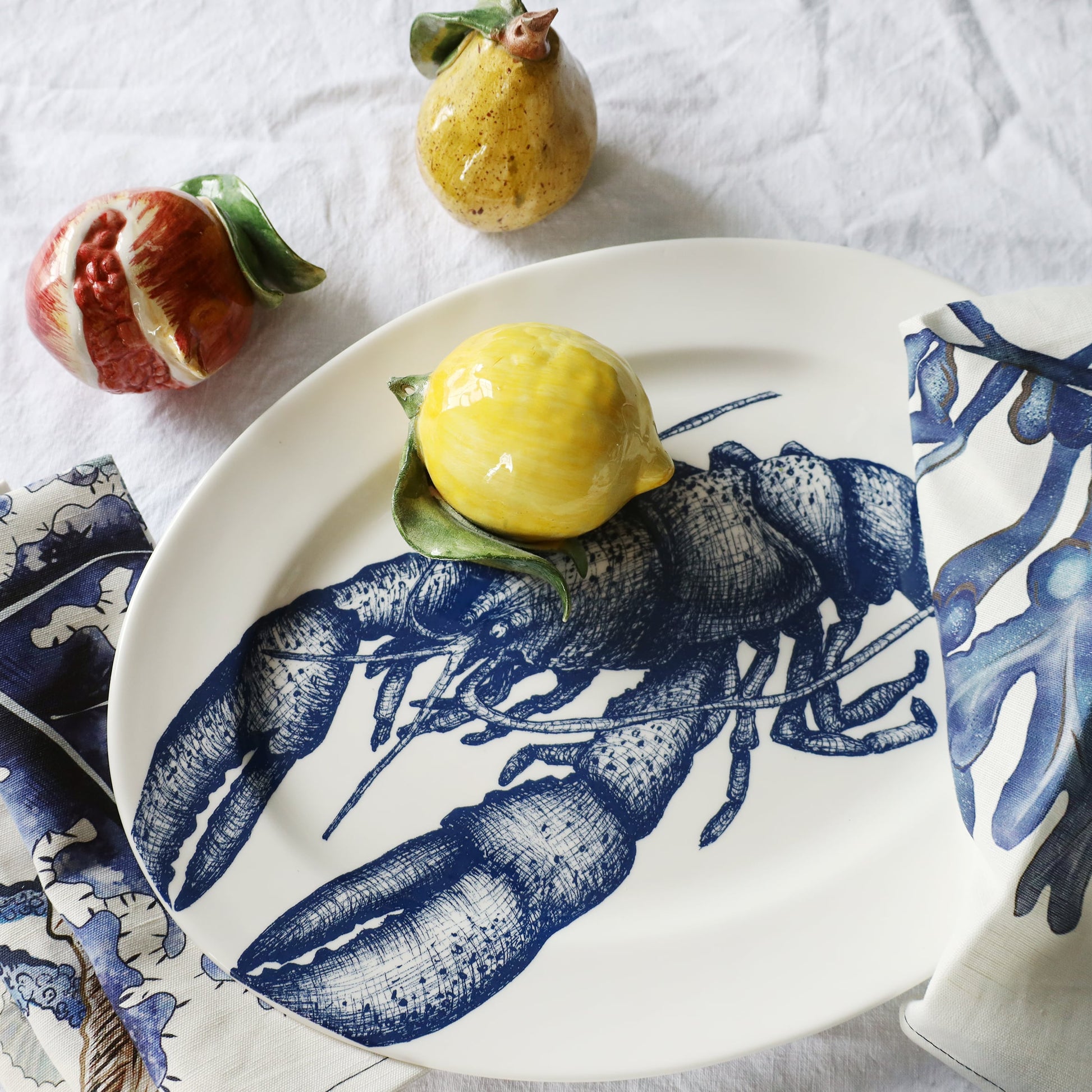 Decorative plate with blue shell design holding a yellow dessert, surrounded by fruits on a white tablecloth.