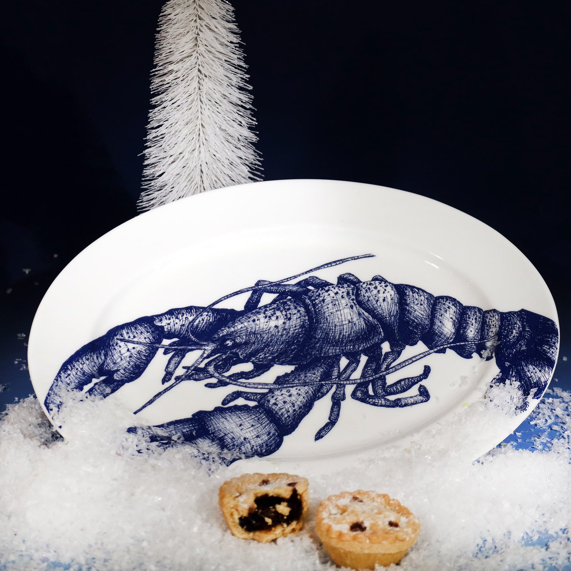 White oval platter featuring a detailed navy blue lobster illustration, styled with faux snow, mince pies, and a white Christmas tree for a festive coastal scene.